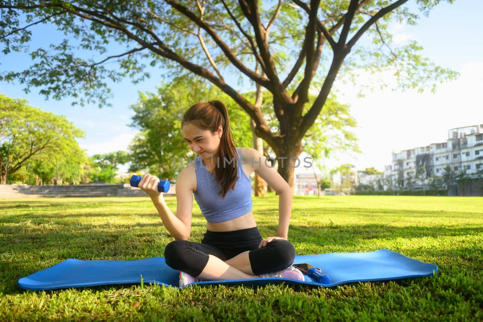 Female fitness enthusiast lifting a dumbbell on mat in a tranquil urban park.