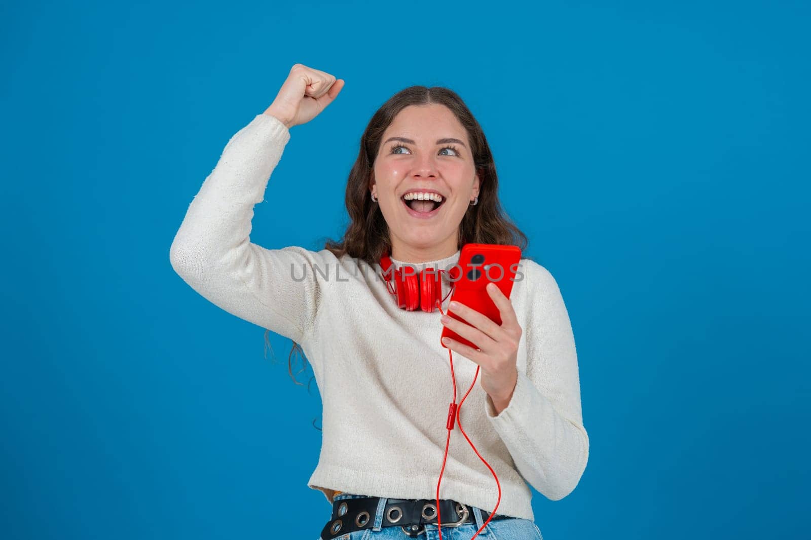 Happy young woman wearing red headphones around her neck, clenching her fist in excitement while looking at her smartphone. Her joyful reaction stands out against the blue background.