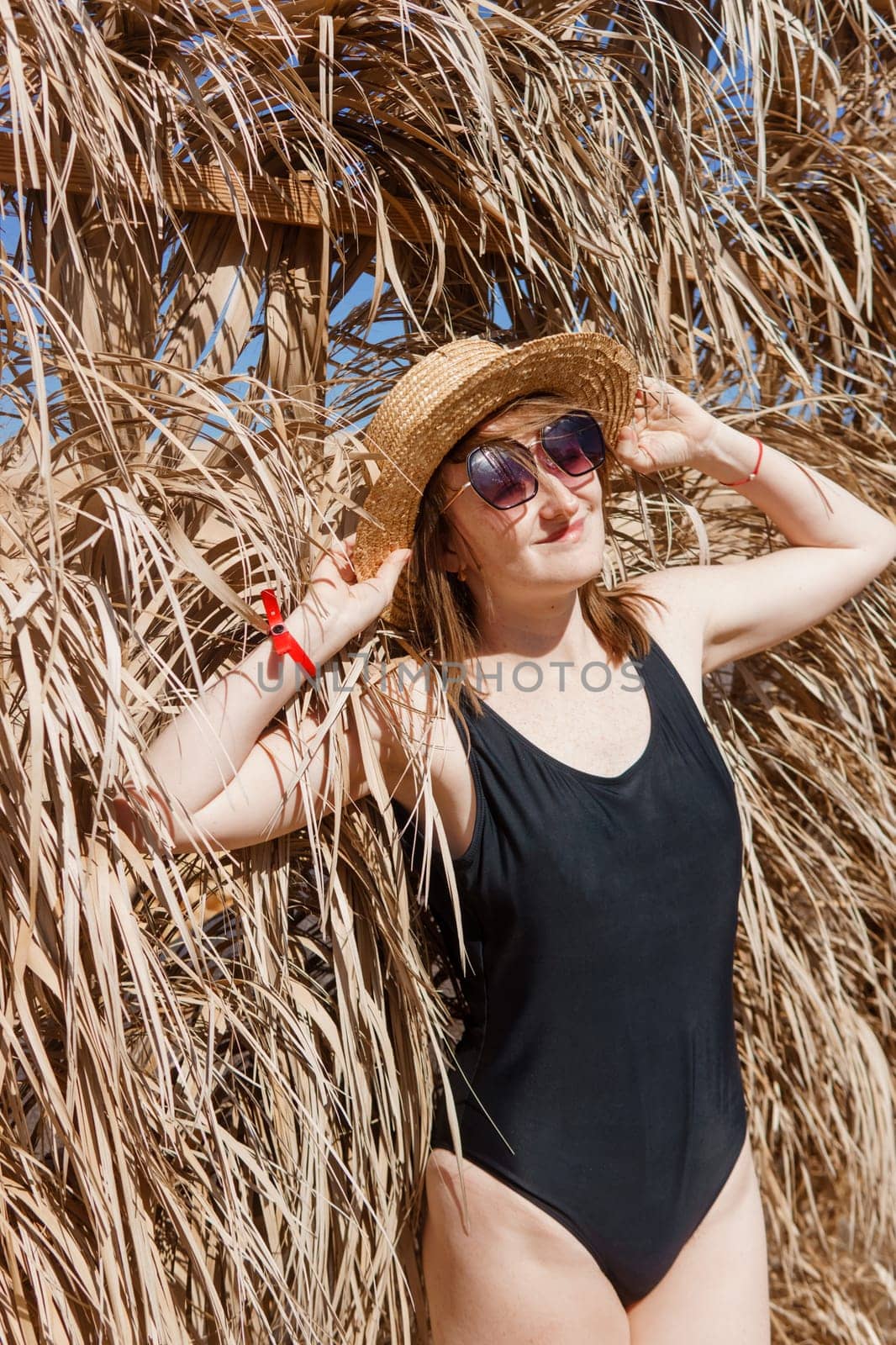 Woman on vacation near straw umbrella on beach. summer holiday concept. woman near palm leaves. person in a one piece swimsuit and sun hat