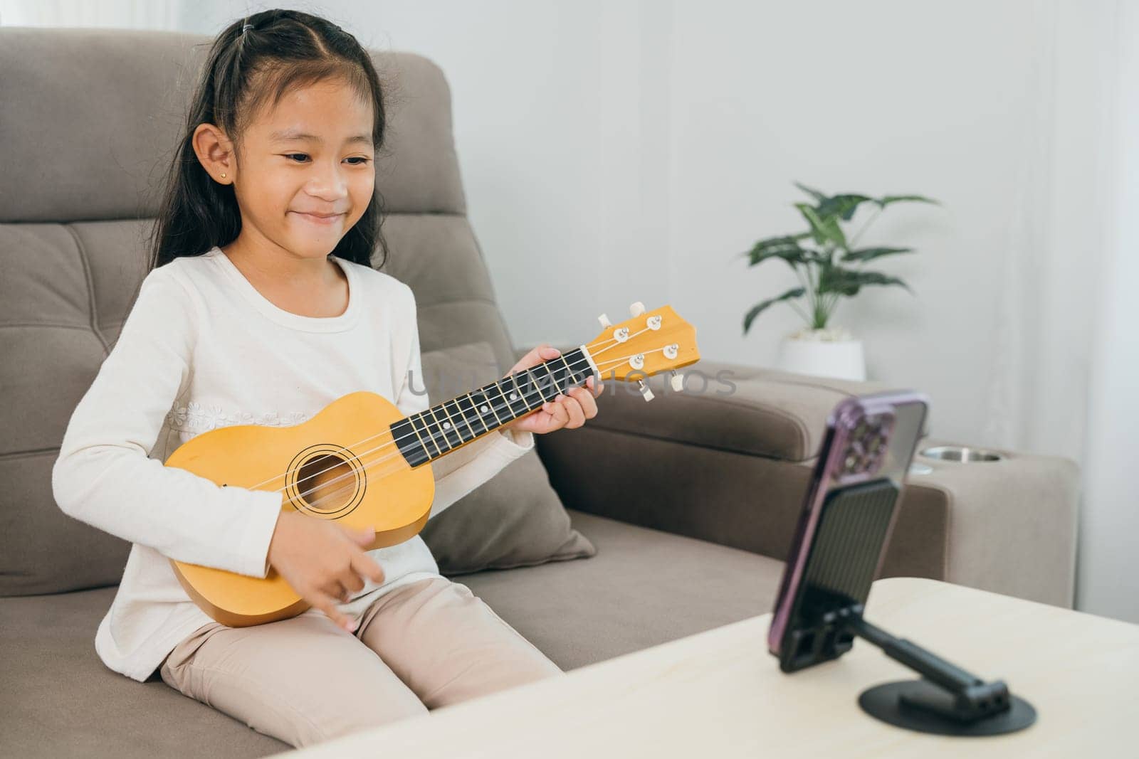 A young girl sits on a couch playing a ukulele, with a smartphone on a stand streaming her online music lesson. The scene highlights creative learning at home with modern digital tools.