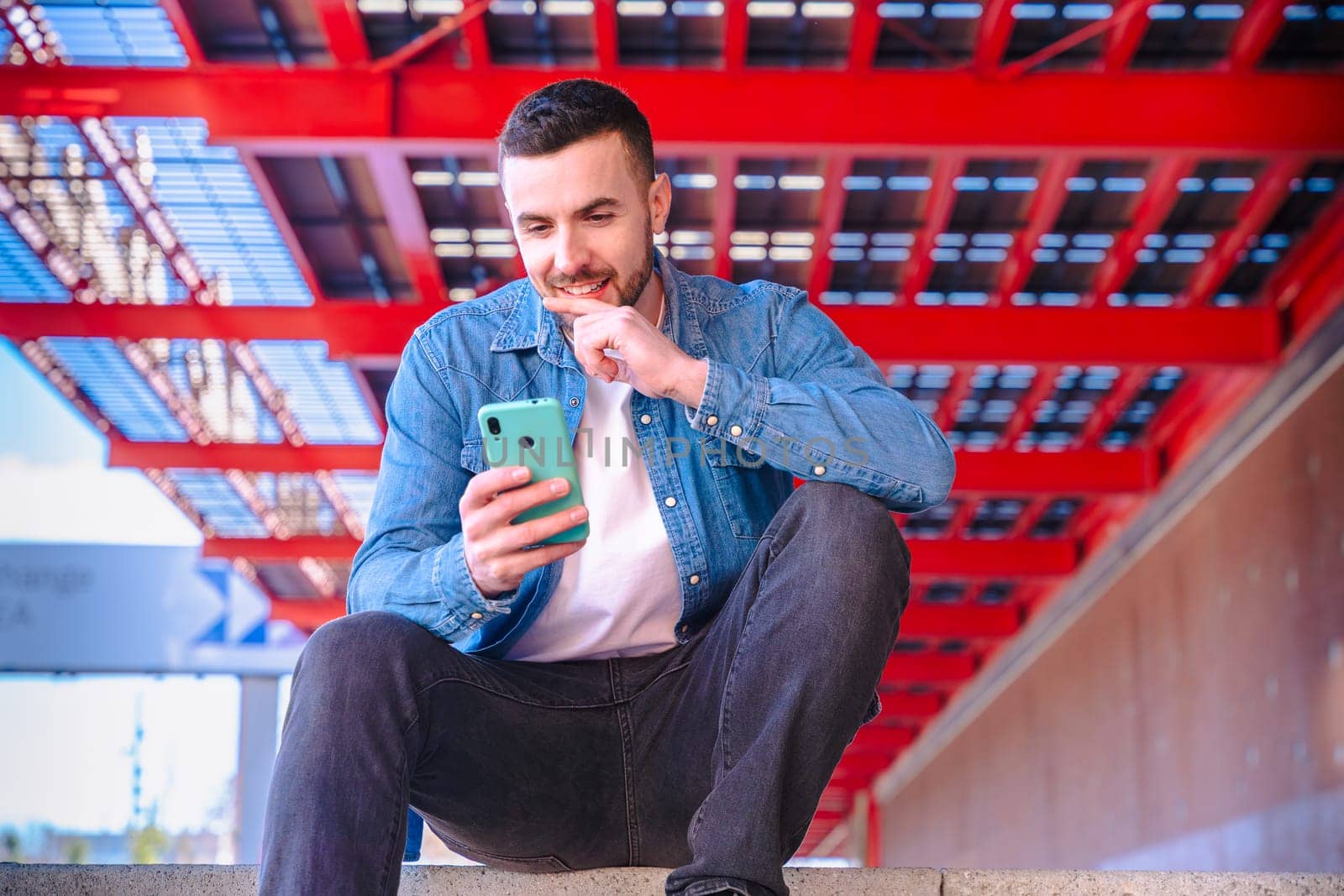 Happy man in denim jacket reacts with excitement while looking at his smartphone. Bright red architectural background in urban setting. Candid expression and joyful moment captured outdoors.