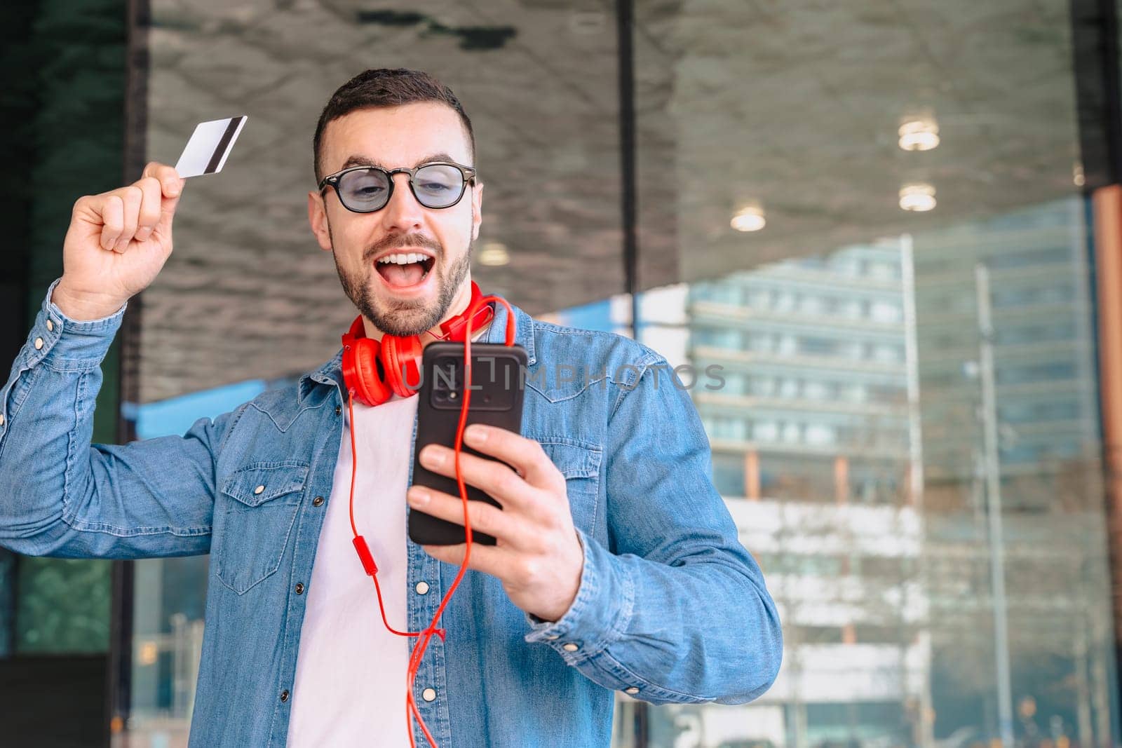 Happy man celebrating a successful online purchase while holding a credit card and smartphone. He smiles with excitement, wearing headphones and sunglasses in a modern urban environment.