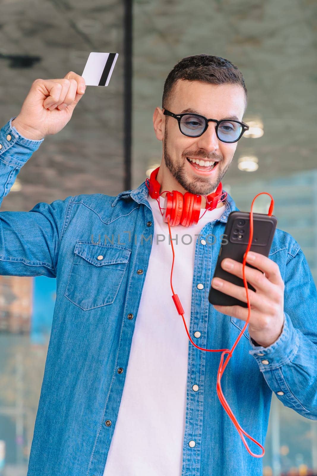 Happy man celebrating a successful online purchase while holding a credit card and smartphone. He smiles with excitement, wearing headphones and sunglasses in a modern urban environment. Vertical