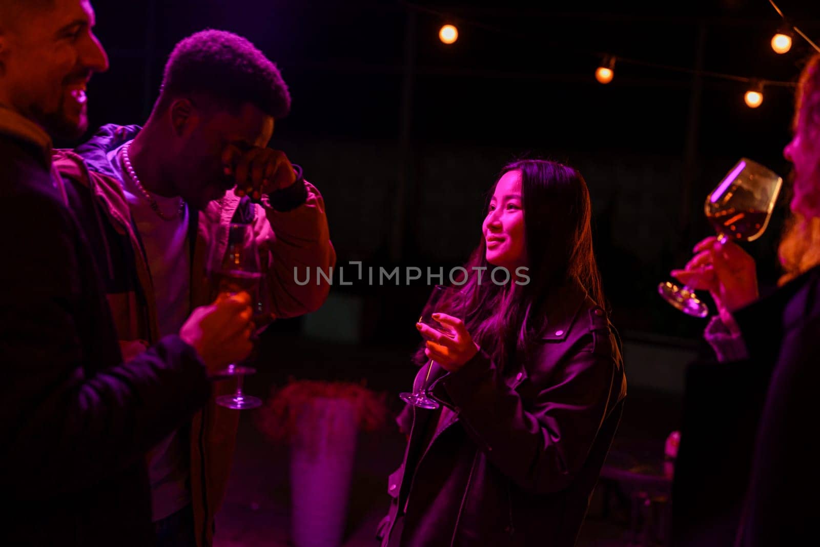 Four friends share laughter and drinks at a lively outdoor party, illuminated by colorful string lights, creating a warm atmosphere during the night.