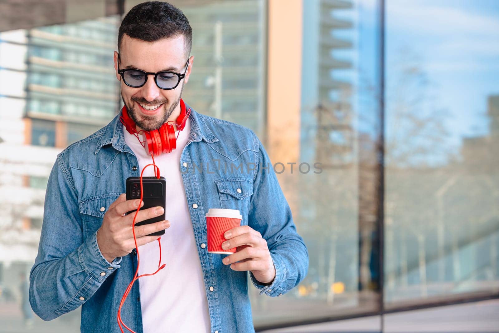 Young man with red headphones enjoying coffee while using his smartphone in a city environment. He smiles while reading or texting, standing outside modern glass buildings in a casual moment.
