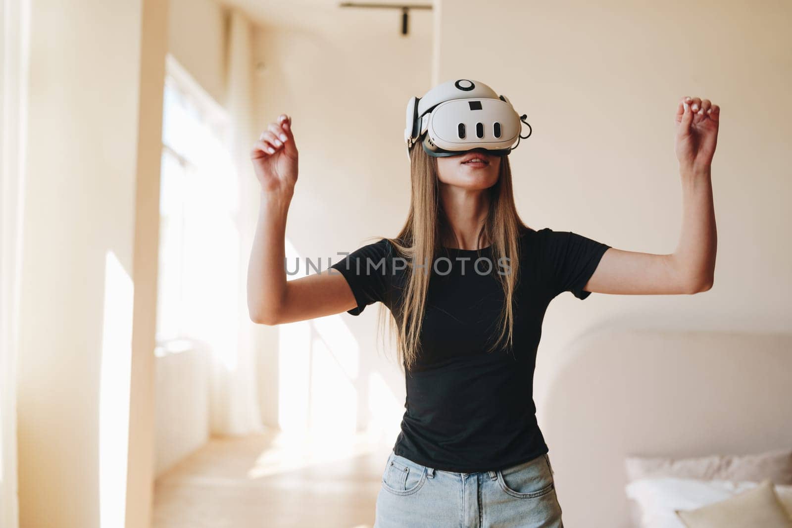 A young woman stands in a bright room wearing a virtual reality headset. She appears immersed in an interactive experience, moving her arms as if engaging with a digital landscape.