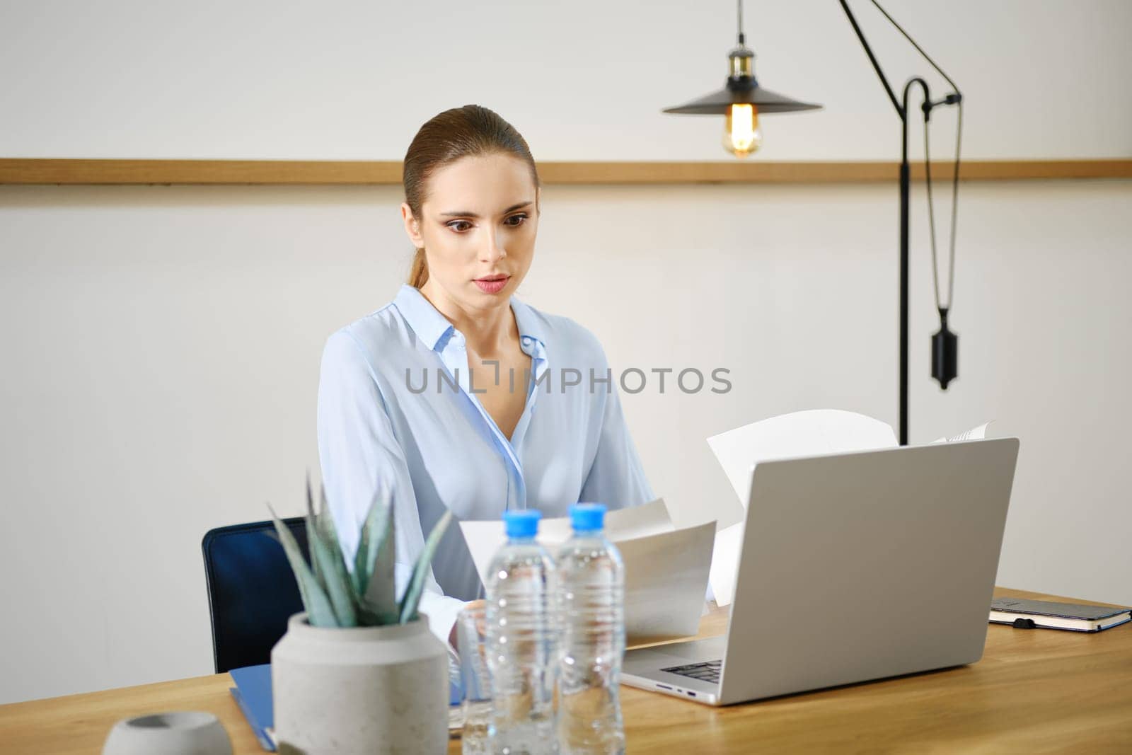 A woman in a light blue blouse is intently reviewing papers at a modern desk. She is surrounded by a laptop, water bottles, and a small plant, all set in a bright office space with minimal decor.