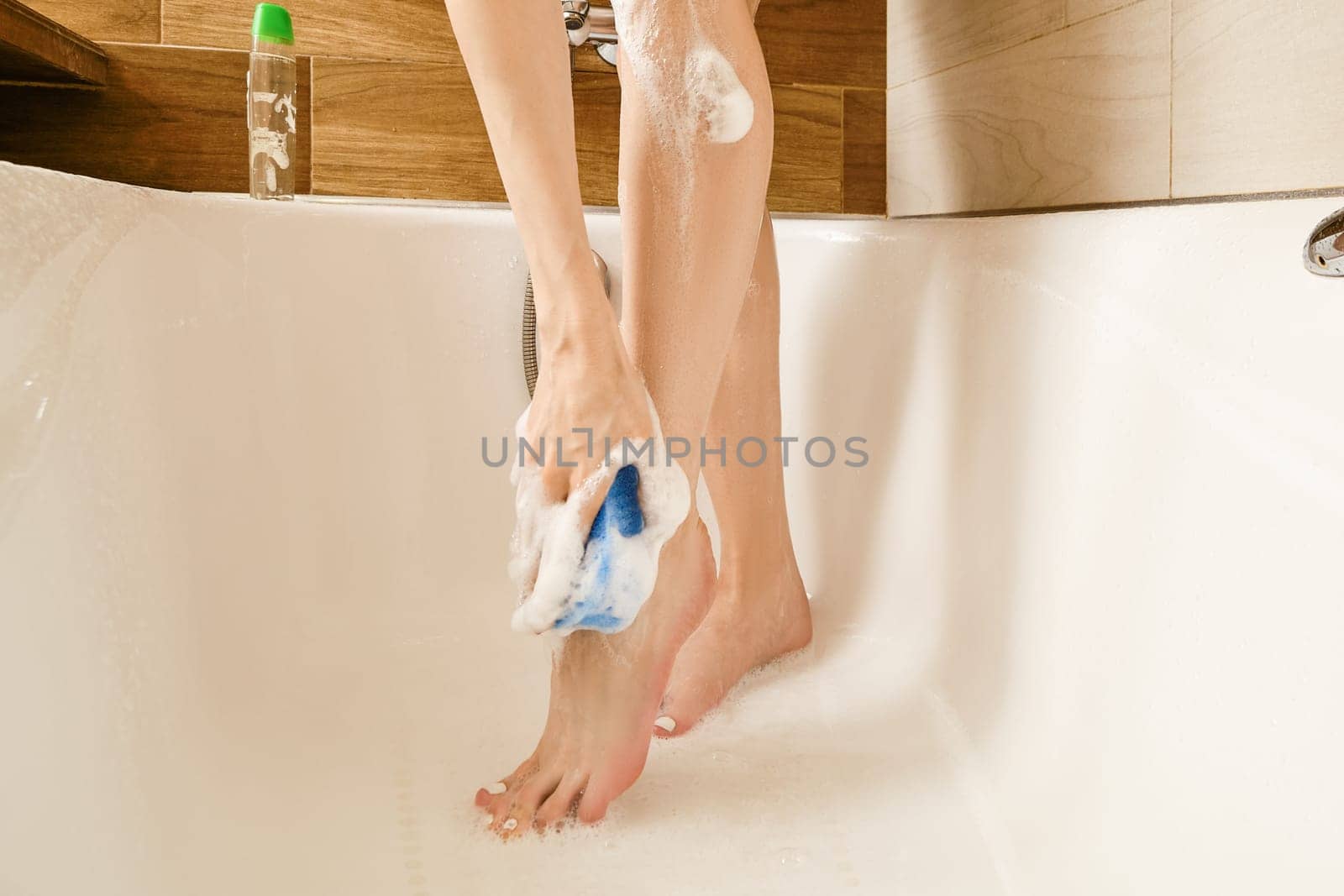 Unrecognizable woman is using a blue sponge to wash her legs, and soap suds are visible on her skin