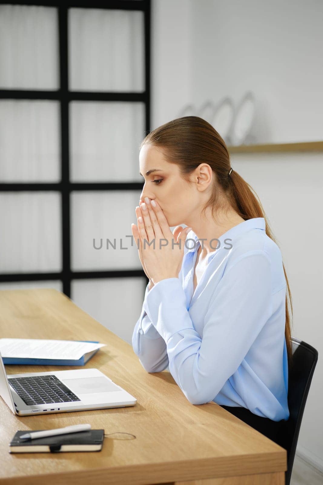 A business woman sits at her desk with a laptop, hands clasped in front of her mouth, visibly deep in thought and reflection