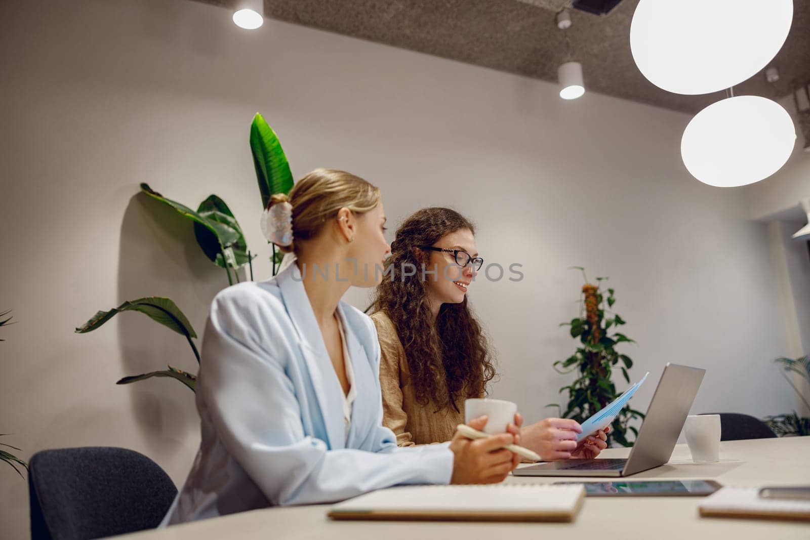 Two professional women collaborate at a stylish office table, surrounded by plants and modern technology