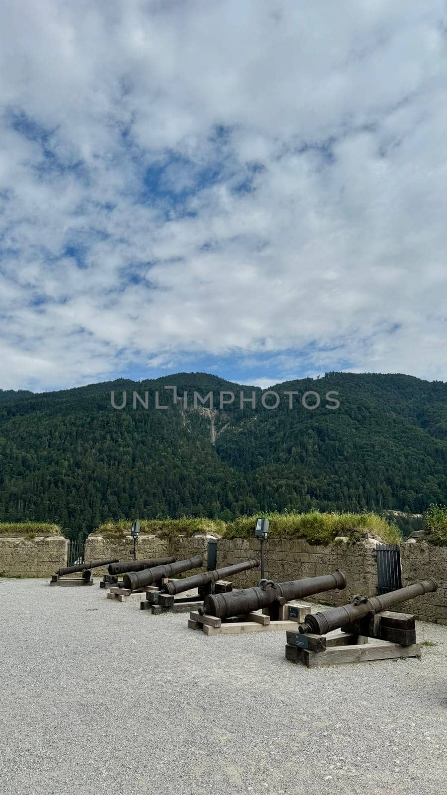 05.08.2024 Austria, Tyrol: Old cannons at Kufstein Fortress with mountain view. Rusty historic cannons displayed on a gravel platform. Set against a backdrop of green mountains and a cloudy sky.