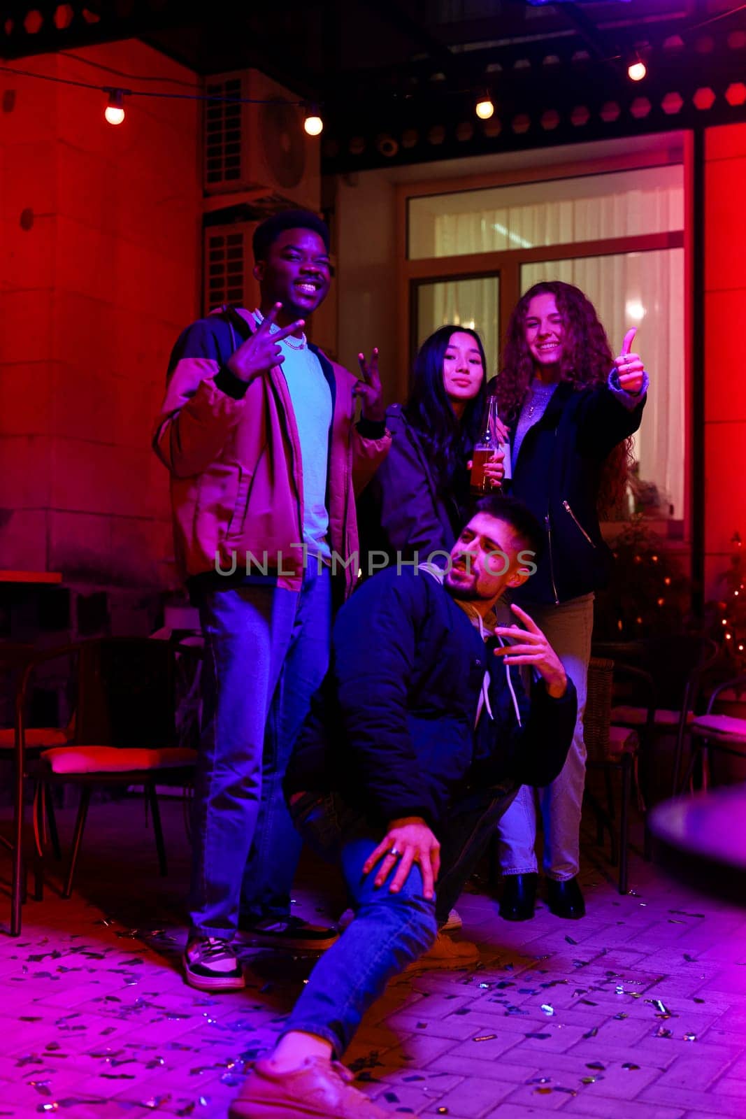Four friends are joyfully posing with drinks and sparklers at a vibrant outdoor venue decorated with colorful lights during a nighttime celebration.