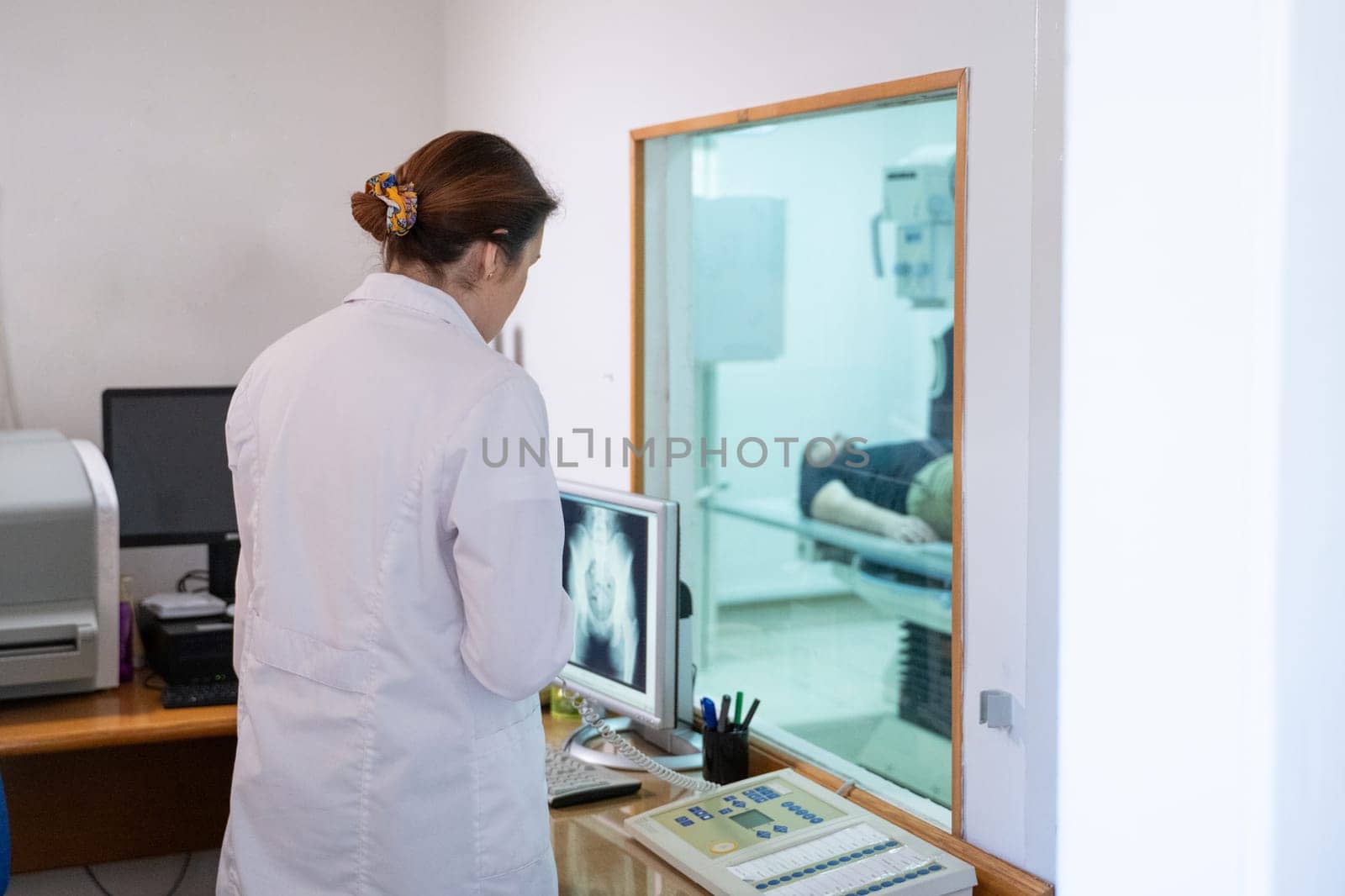 A radiologist in a white lab coat sitting at a workstation while analyzing an X-ray image of a patients hip in a medical imaging room.