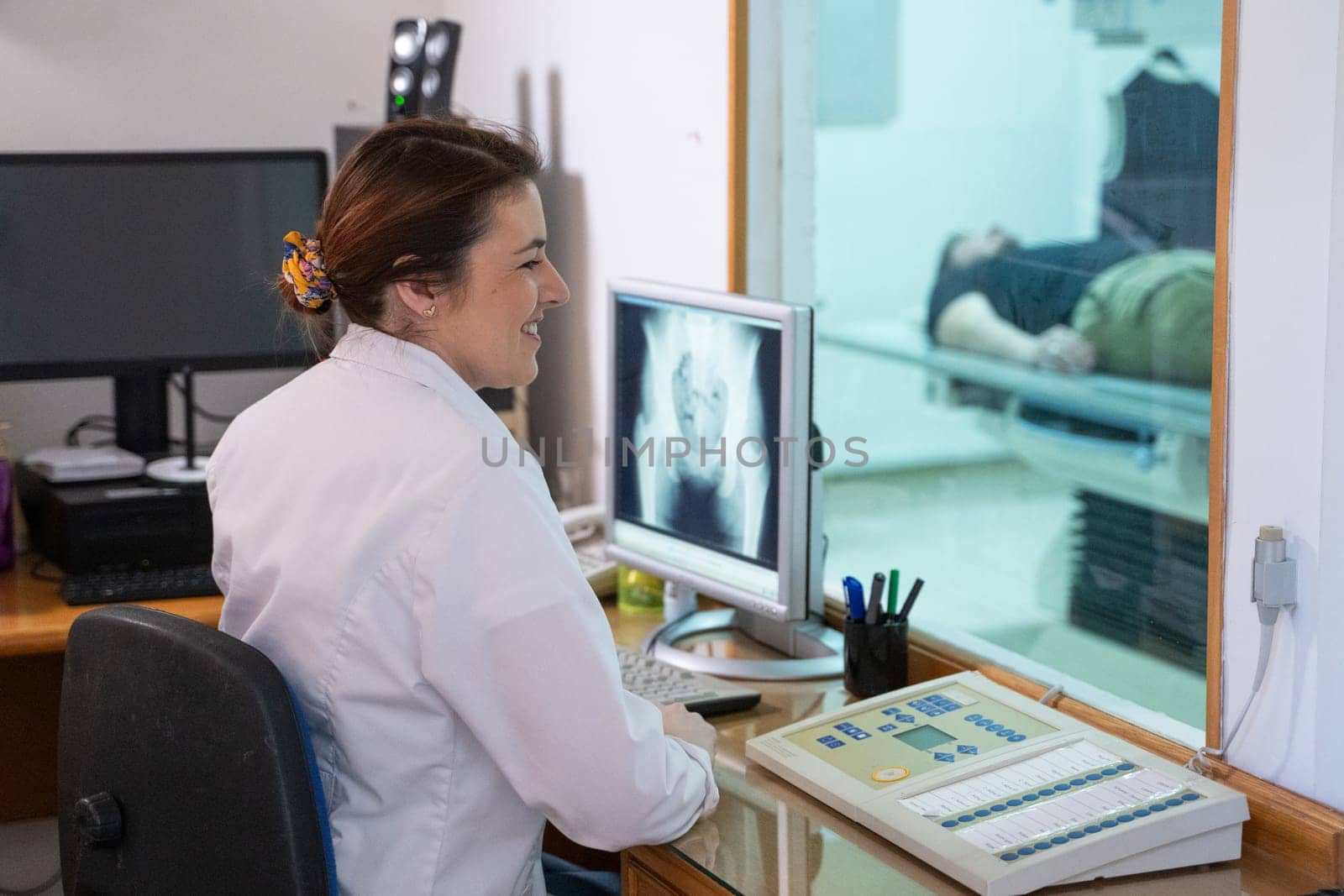 A radiologist in a white lab coat sitting at a workstation while analyzing an X-ray image of a patients hip in a medical imaging room.