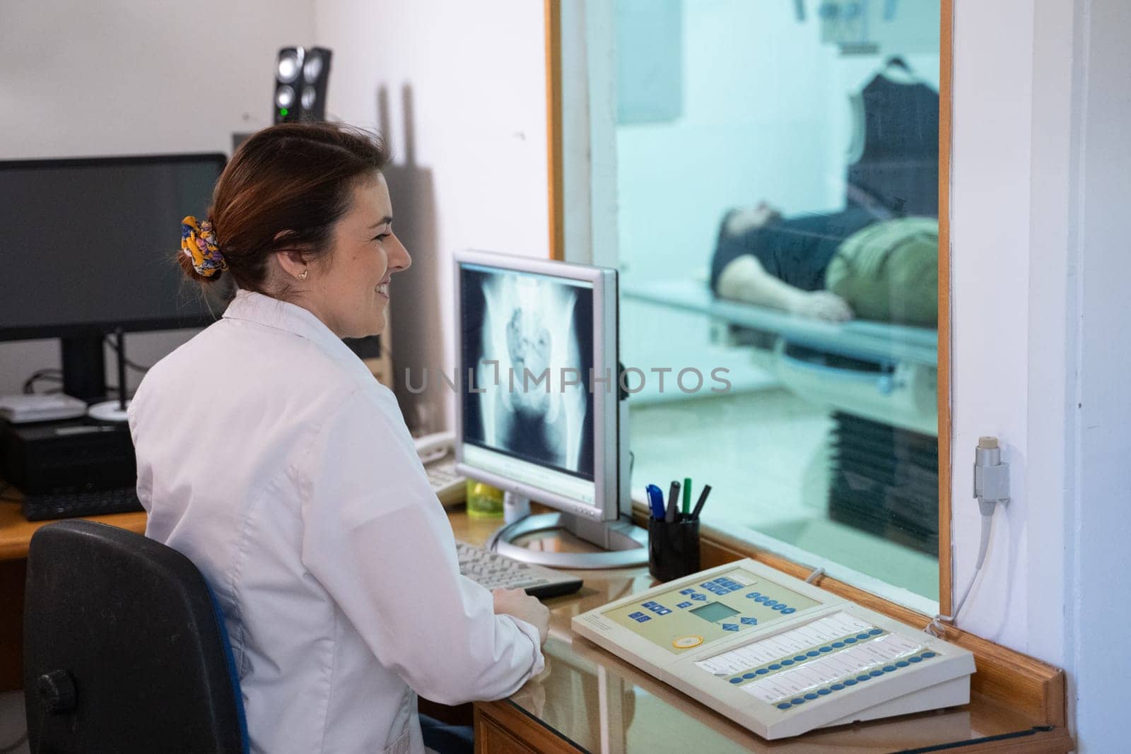 A radiologist in a white lab coat sitting at a workstation while analyzing an X-ray image of a patients hip in a medical imaging room.