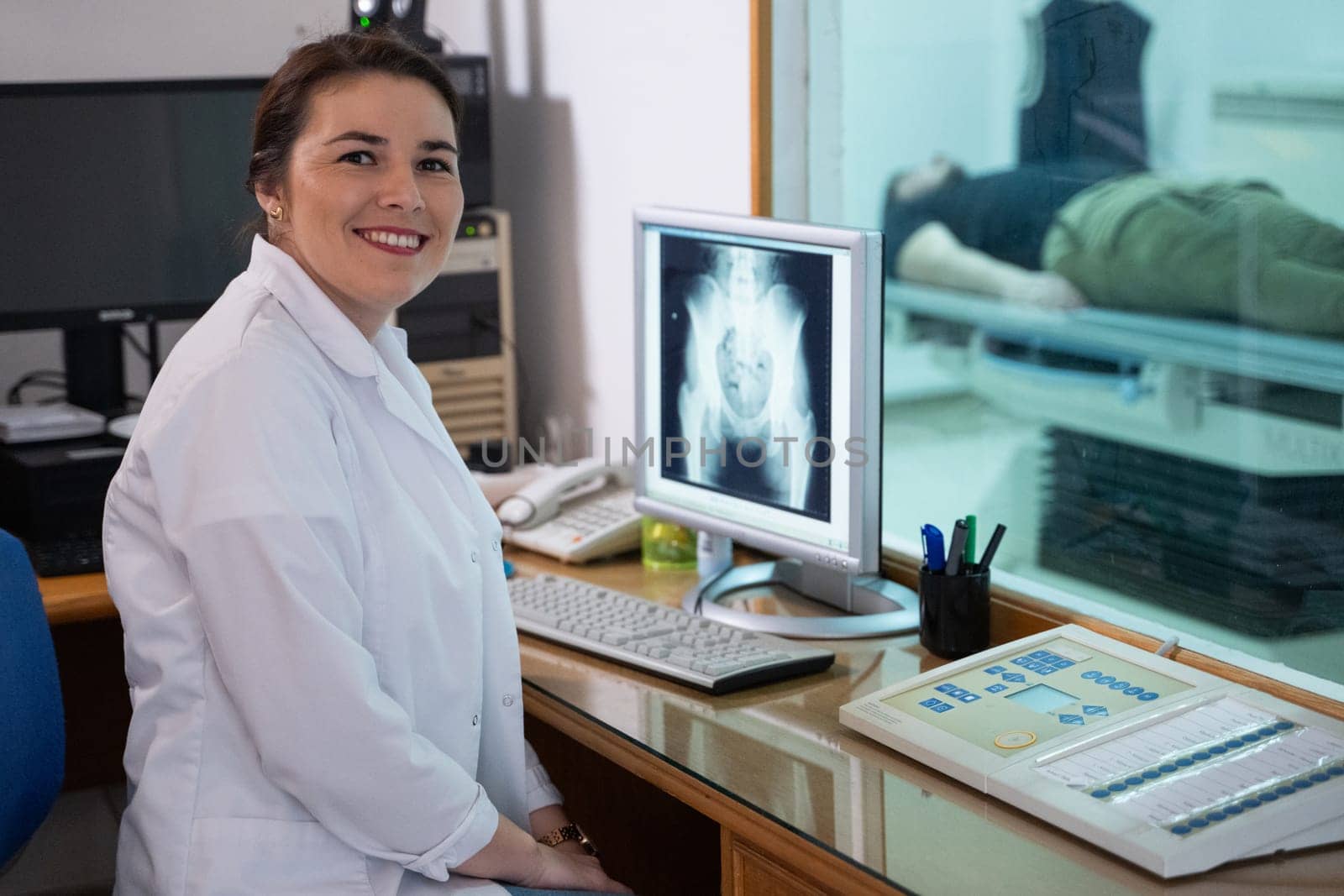 A radiologist in a white lab coat sitting at a workstation while analyzing an X-ray image of a patients hip in a medical imaging room.