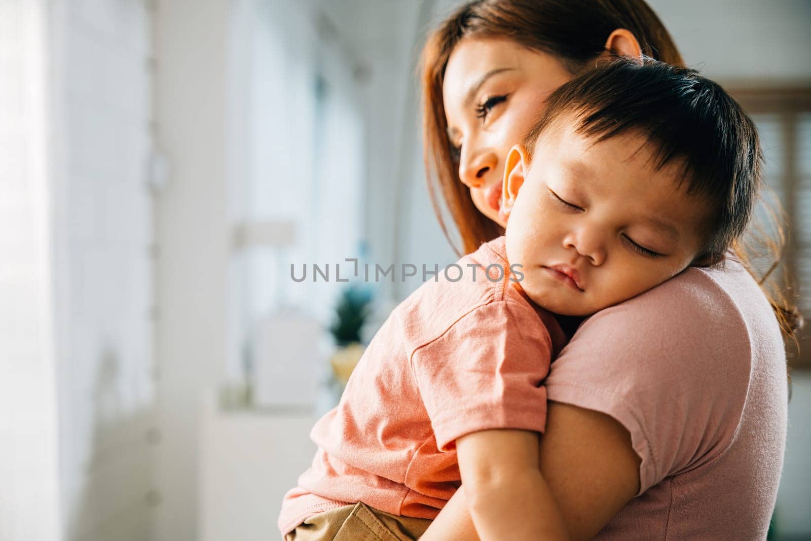 In the warmth of their home an Asian mom gently embraces her small sleeping newborn cherishing a precious family moment filled with the wonder of new life and the carefree moments of childhood.