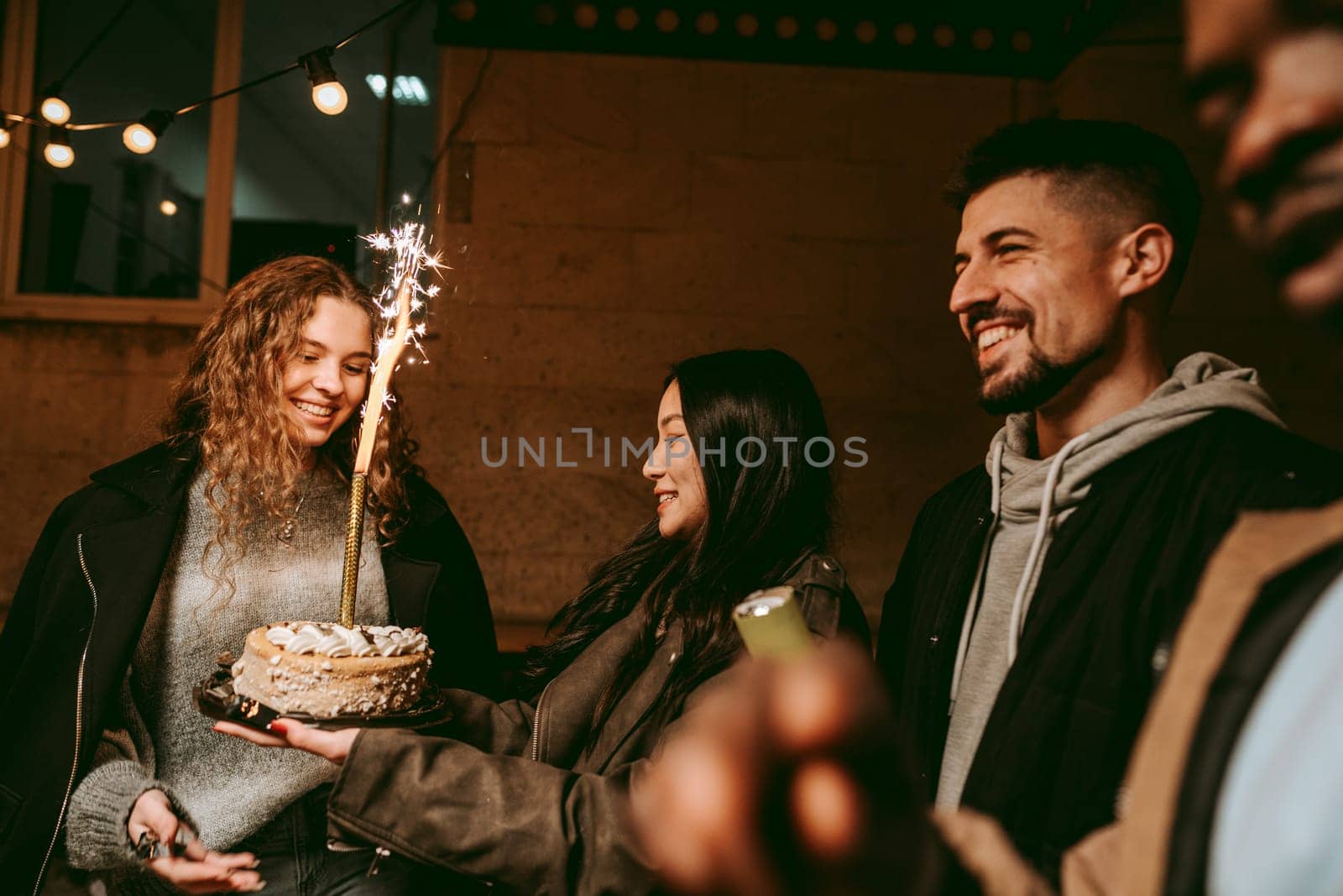 A group of friends joyfully celebrates a birthday at night. One friend holds a beautifully decorated cake with a sparkler, while others share smiles and laughter, enjoying the festive atmosphere.