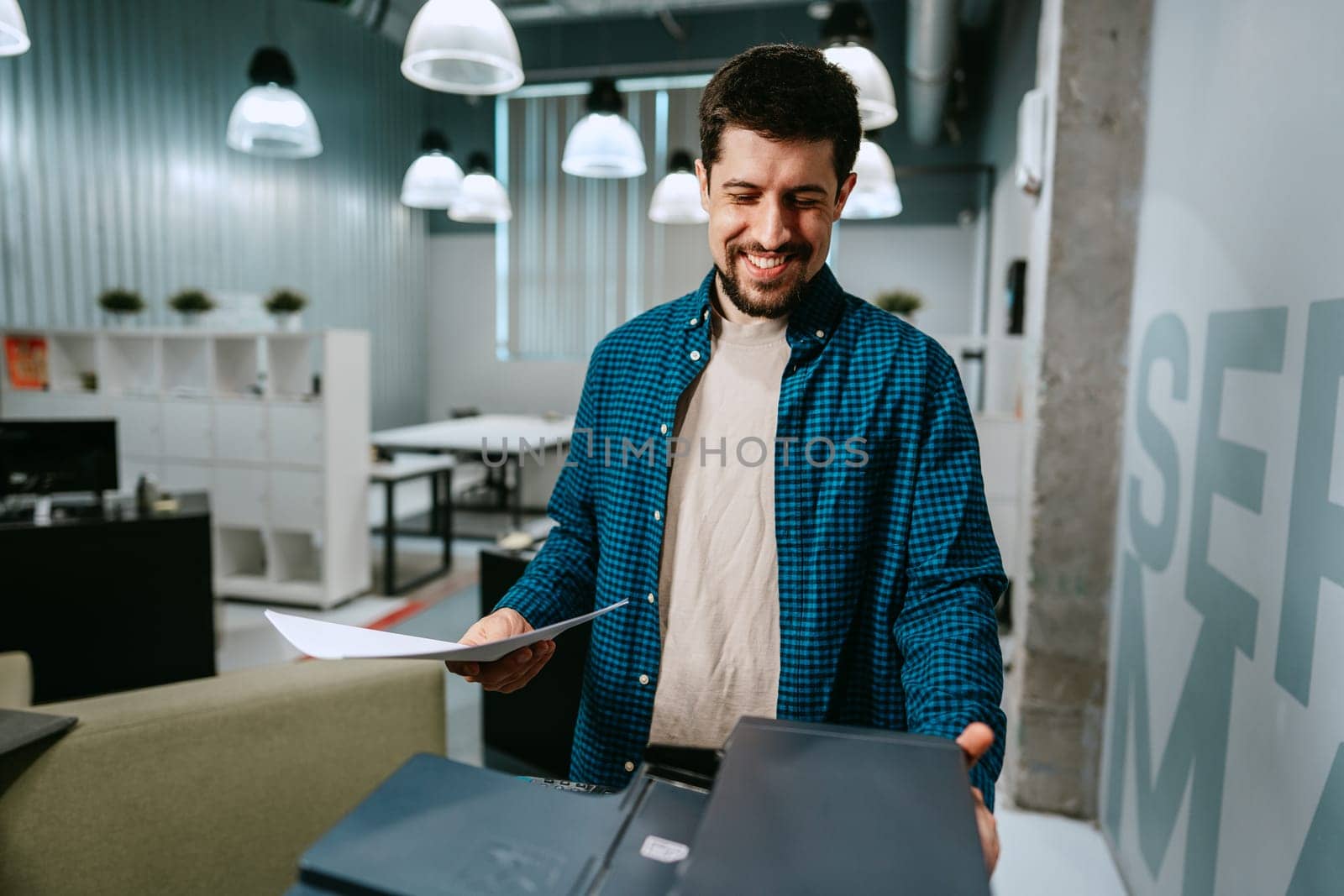 A man stands in a contemporary office, smiling as he examines papers. The space features modern decor, bright lighting, and workstations in the background.