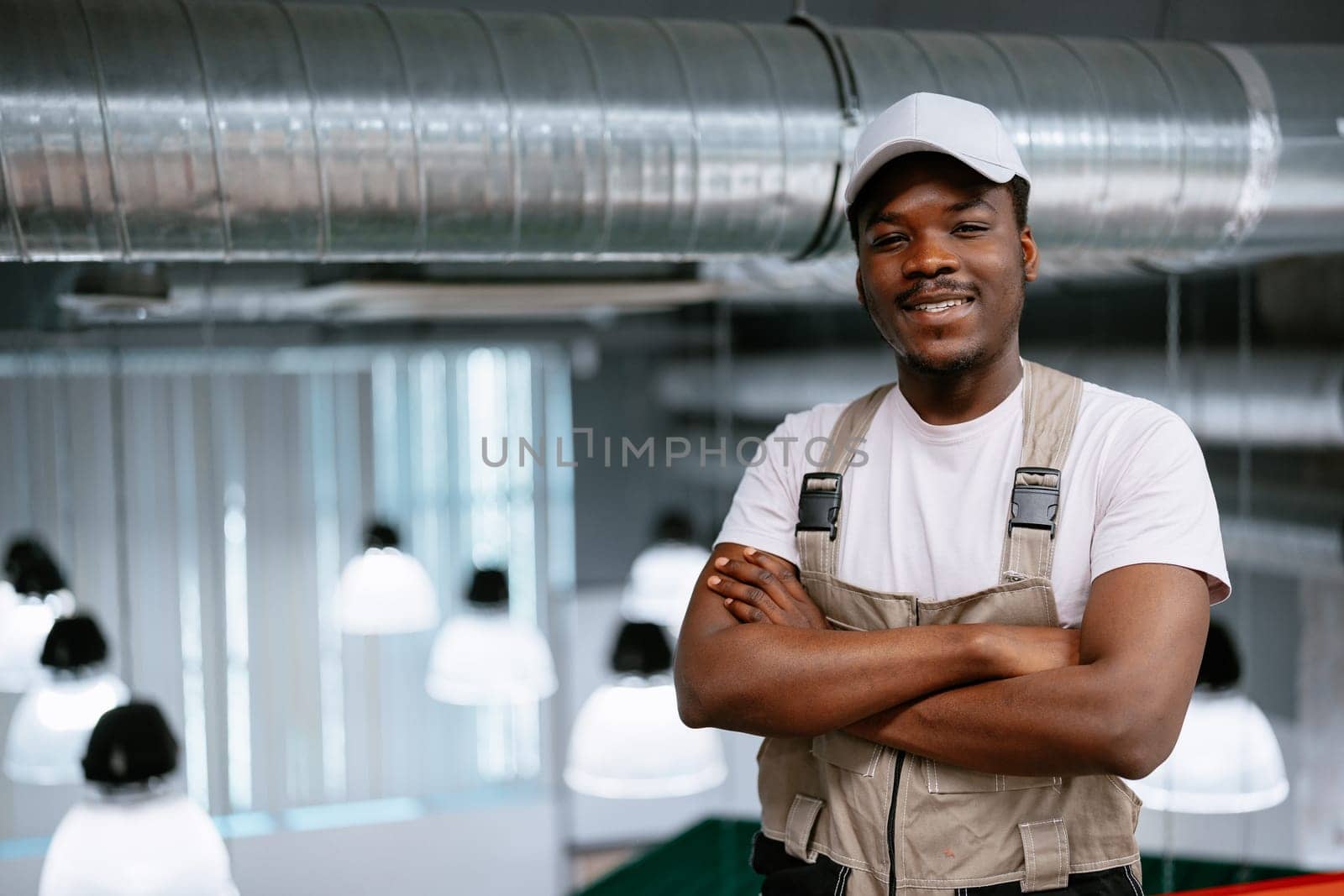 A young man in a casual outfit poses with crossed arms in a contemporary work environment featuring industrial design. The background highlights distinctive light fixtures and exposed ducts.
