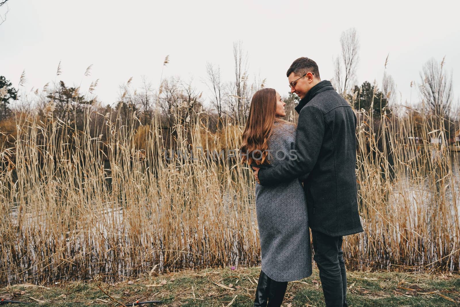 A couple stands closely by a calm waterway, sharing a warm embrace amidst tall reeds. The tranquil autumn setting creates a cozy atmosphere as they enjoy each other's company.
