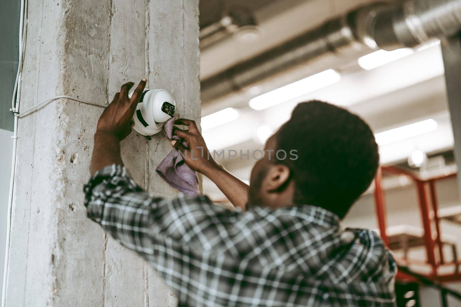 A worker in casual attire is using a cloth to clean a surveillance camera mounted on a concrete pillar, ensuring the equipment functions properly in a modern industrial environment.