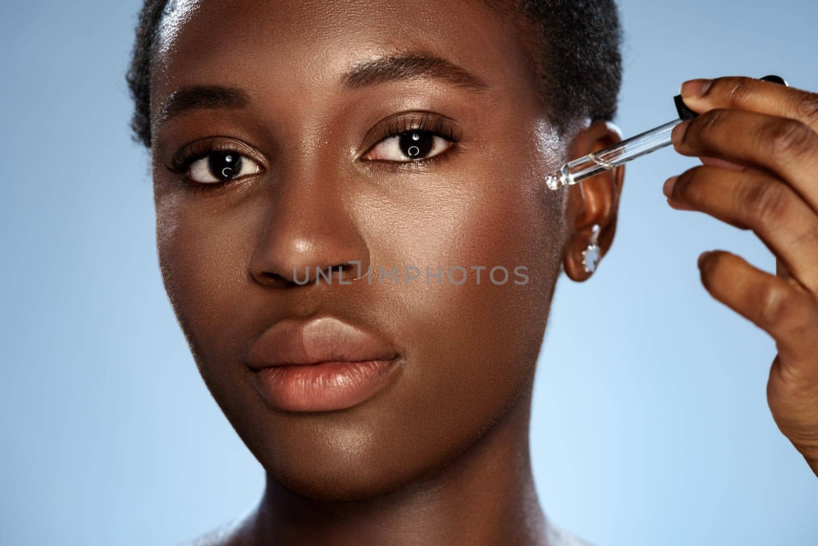 A woman applies a serum to her face using a dropper, showcasing her skincare routine. The setting is bright with a smooth backdrop enhancing her natural beauty.
