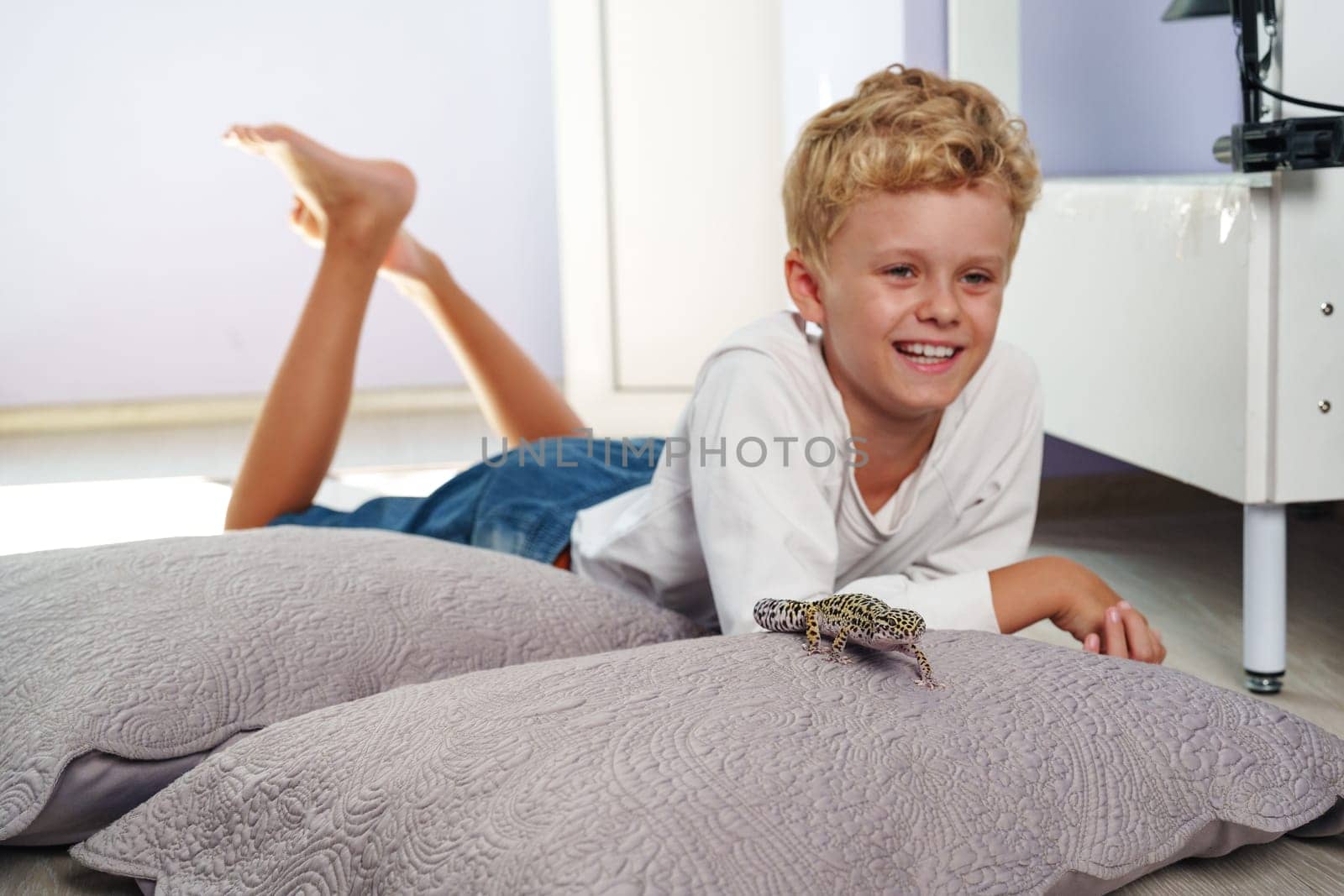 Child lays on large pillows in a cozy room, smiling as he interacts with a gecko. Sunlight brightens the space, showing a relaxed afternoon at home.