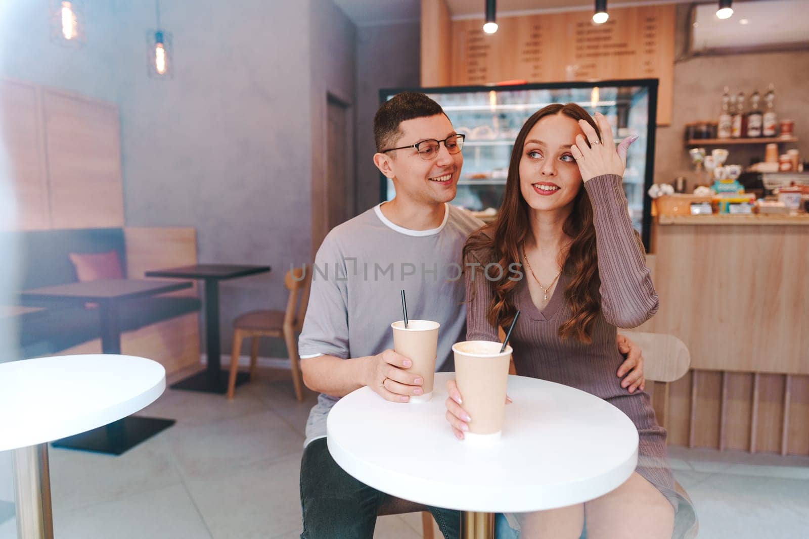 A couple sits close together at a small table in a bright cafe, sipping coffee from paper cups. They are smiling and engaging in a lighthearted conversation, creating a warm atmosphere.