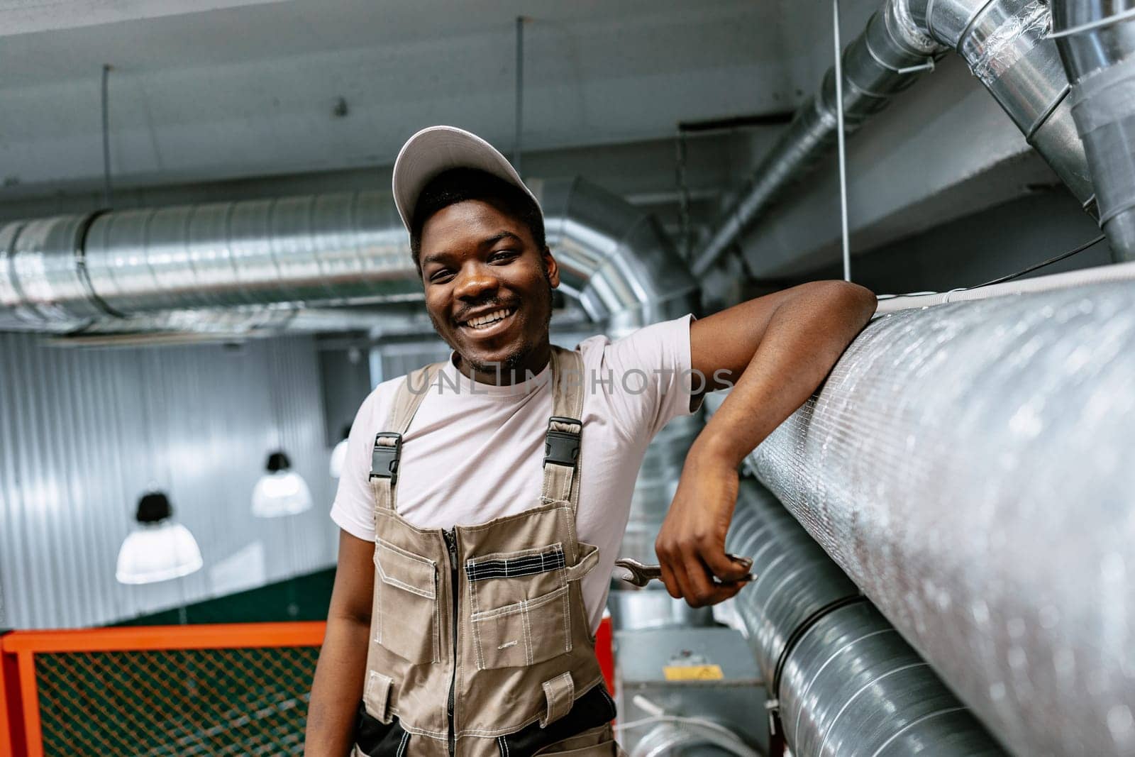 A cheerful worker stands confidently on a platform in an industrial setting, wearing safety gear and a cap. The bright environment showcases pipes and overhead lighting.