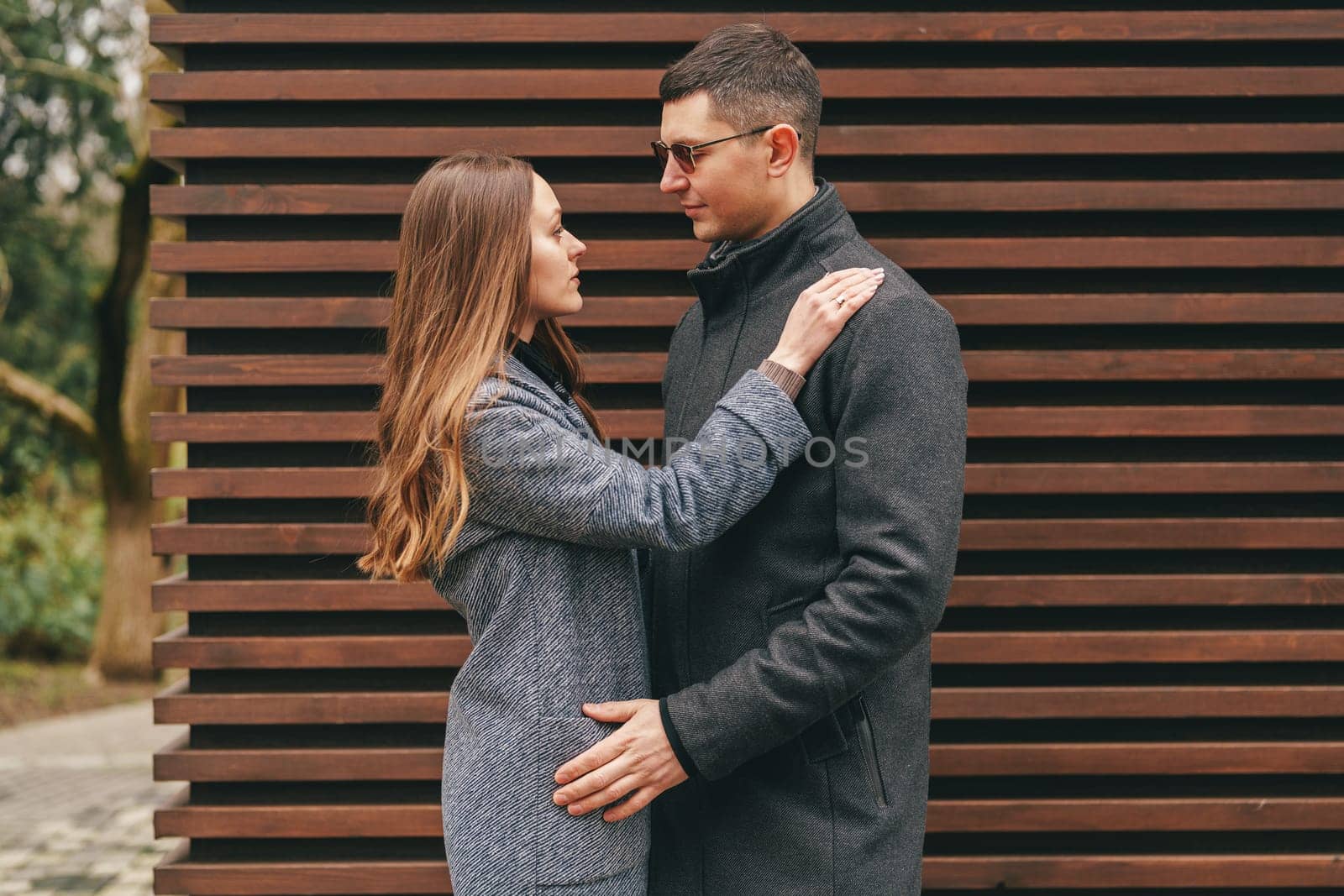 A couple stands close together in a contemporary outdoor setting, sharing a moment of affection. The woman gazes lovingly at the man, who gently rests his hand on her back.