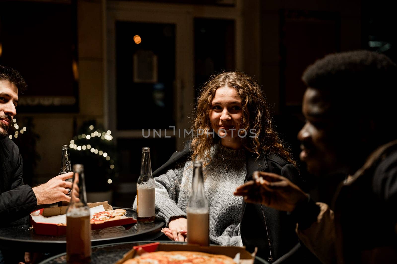 Three friends share laughter and conversation while eating pizza and drinking beverages at an outdoor table. The evening ambiance is warm and inviting.