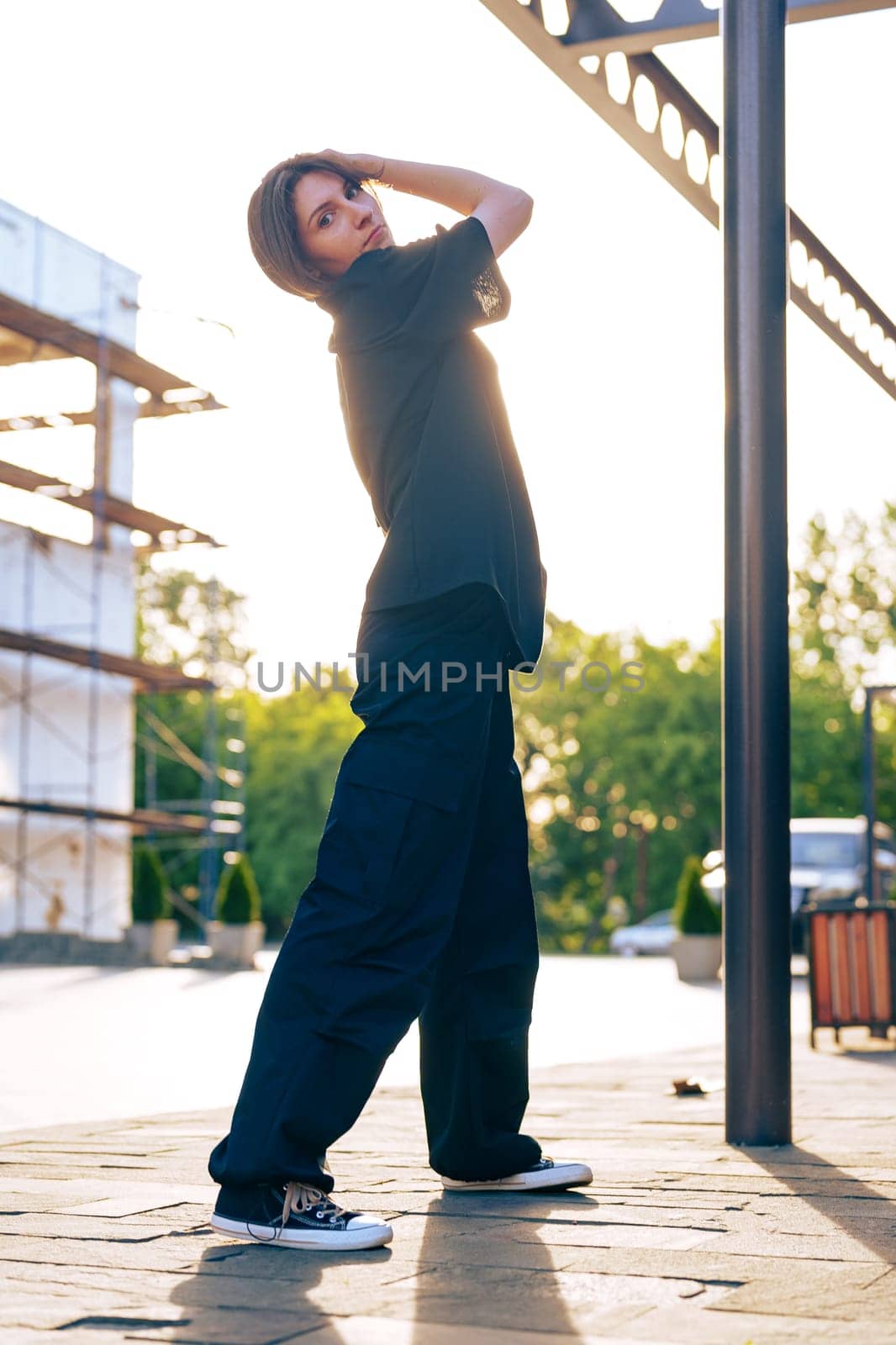 A woman stands confidently outdoors in comfortable attire, with the sunlight casting a warm glow. The background features construction scaffolding and greenery, creating a vibrant atmosphere.