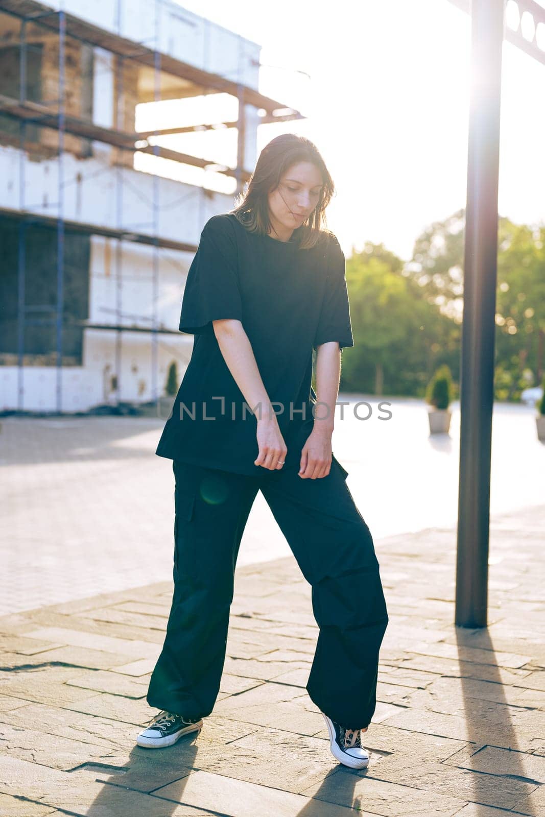 A young woman stands on a cobblestone path in a casual black outfit, surrounded by an urban environment under soft evening light, showcasing her relaxed style.