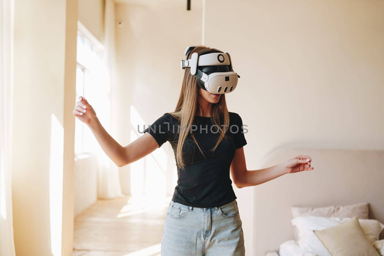 A woman in a casual black t-shirt and jeans is immersed in a virtual reality activity at home. She appears to be interacting with digital content in a bright, sunlit room.