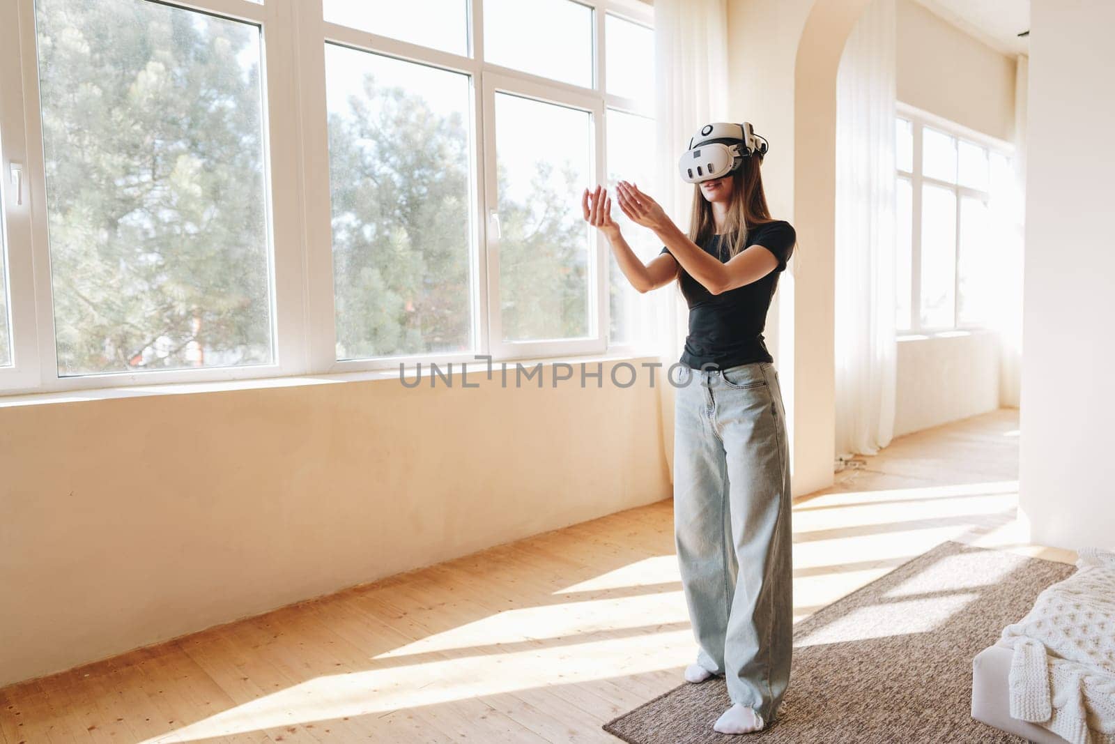 A woman is fully immersed in a virtual reality session, wearing a headset and using hand gestures to interact. The spacious room is filled with natural light, highlighting her concentration.
