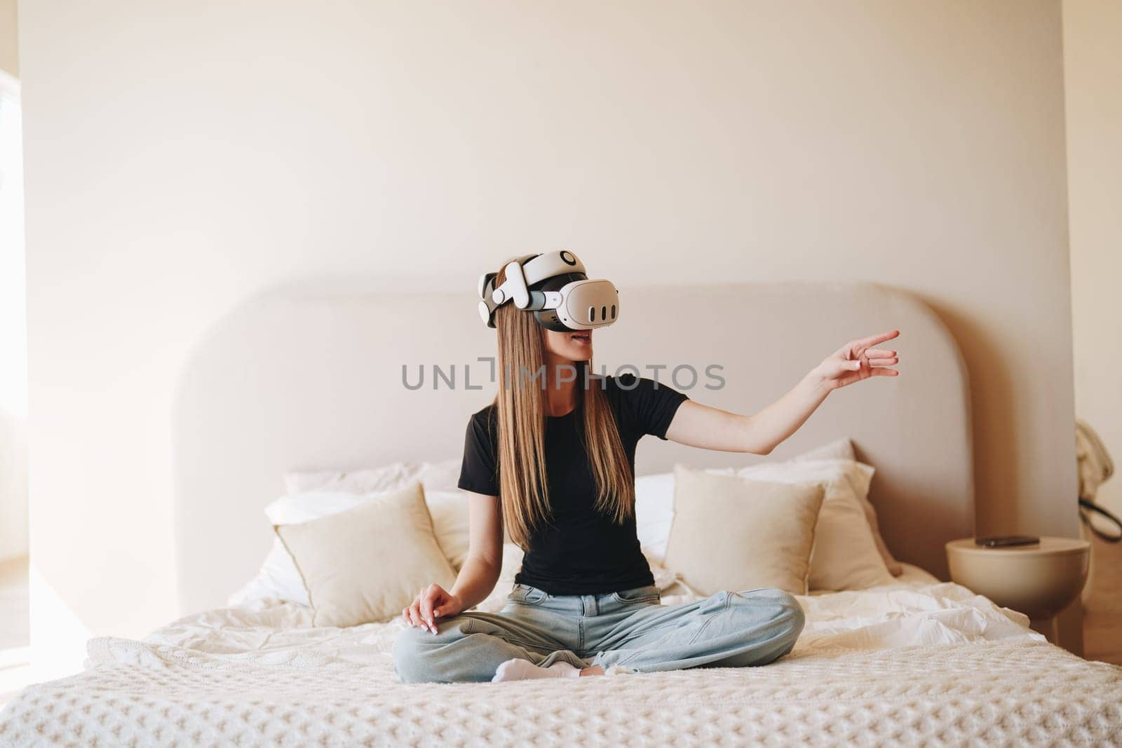 A young woman is seated cross-legged on a bed, wearing a virtual reality headset and fully engaged in the immersive experience.
