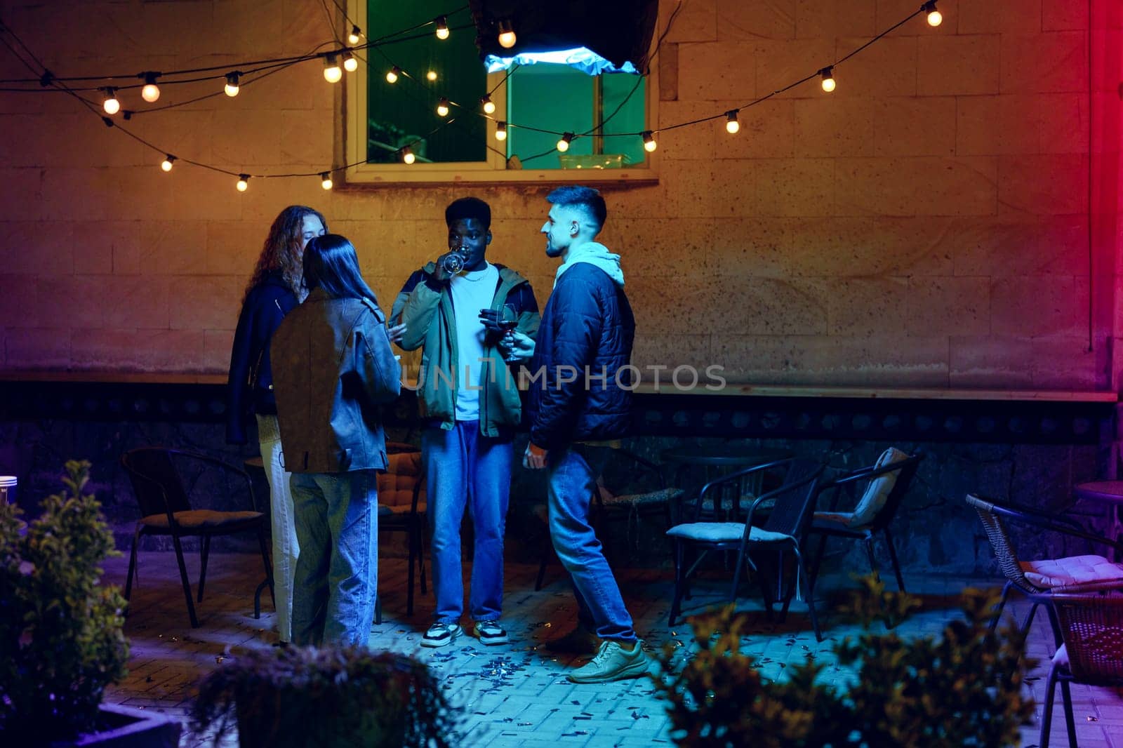 A group of four friends socialize outdoors during the evening, surrounded by string lights. They share drinks and laughter in a cozy, casual ambiance.