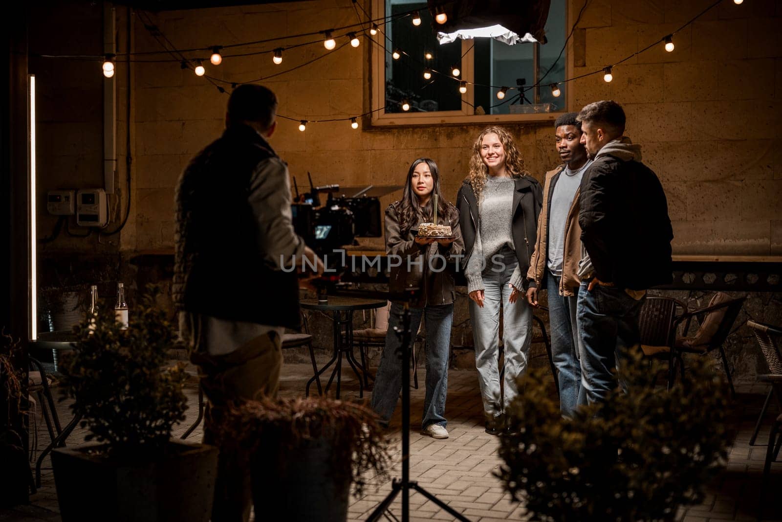 A diverse group of four individuals stands together, posing cheerfully in an outdoor setting lit by string lights. The atmosphere appears relaxed as they engage with a camera operator.