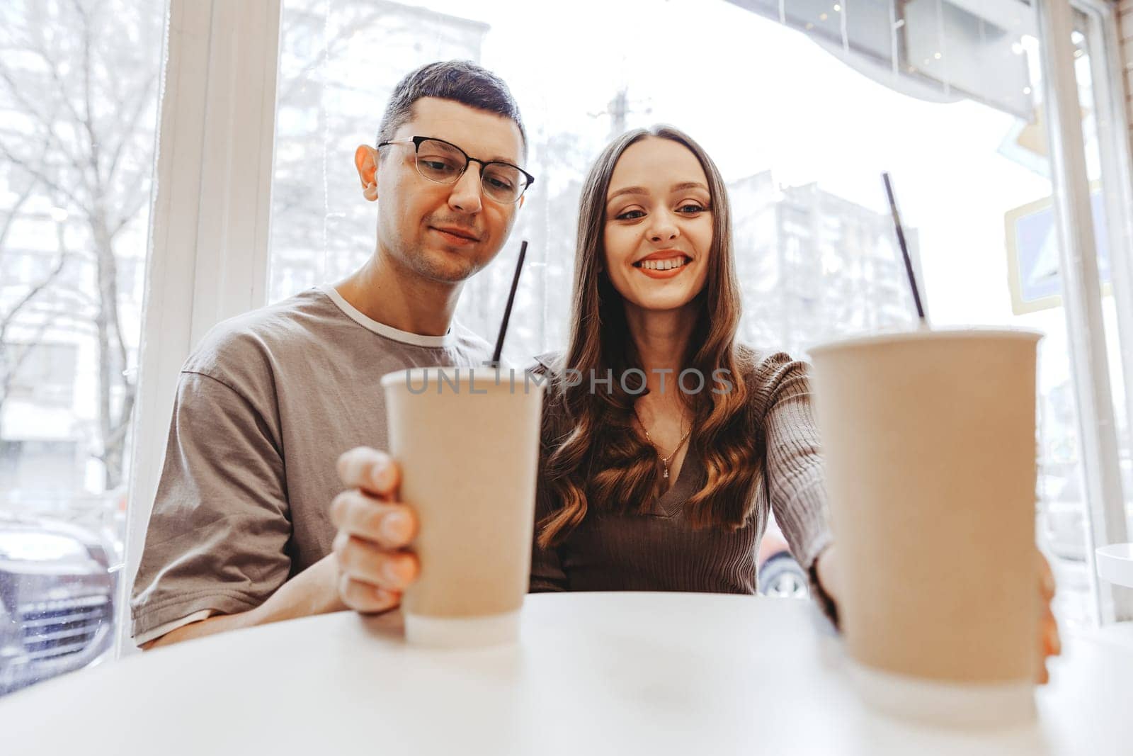 A young couple smiles while sitting at a cafe table, holding disposable cups filled with drinks. The backdrop shows a busy city street through the window.