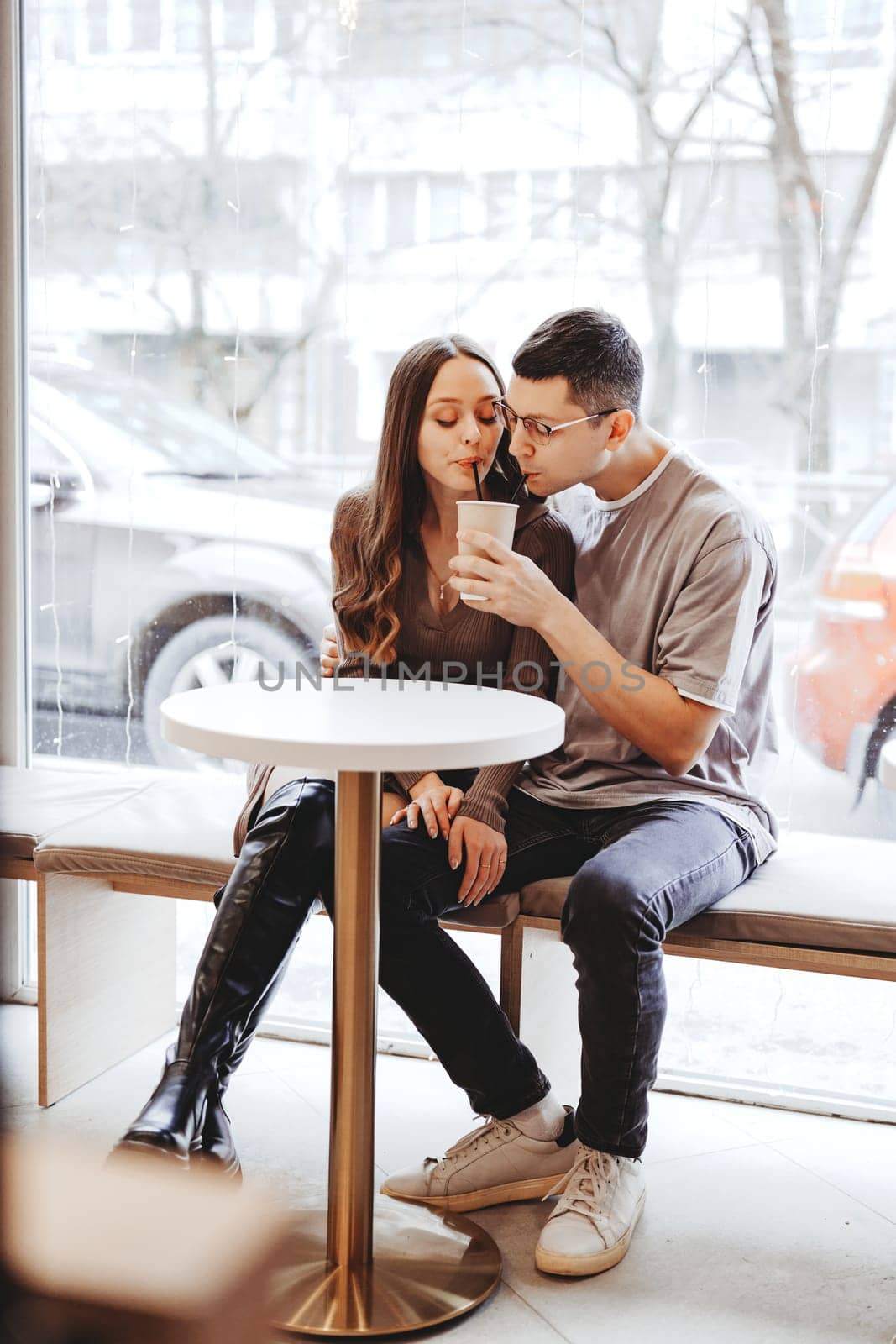 Two individuals share a tender moment in a cafe, sipping warm beverages while snow gently falls outside. They sit closely, creating a warm and intimate atmosphere.