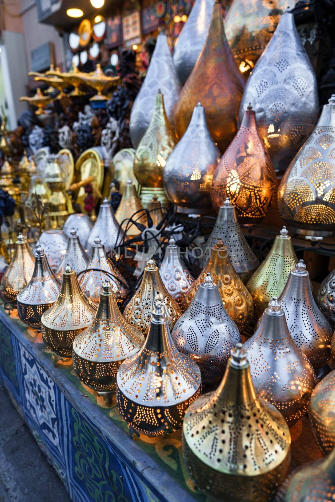 A vibrant market stall displays an array of decorative lamps in gold and silver tones. Intricate patterns adorn the lamps, reflecting the warm early evening light and attracting visitors.