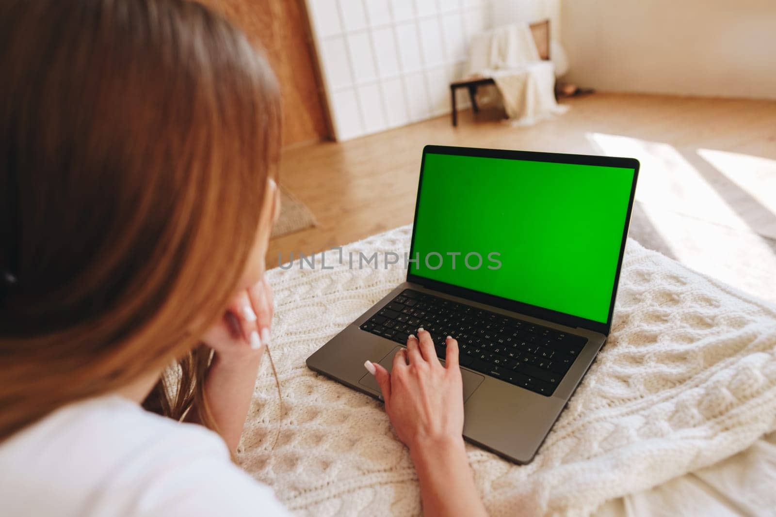 A woman sits on a bed covered with a soft blanket, intently working on a laptop displaying a bright green screen. Sunlight streams through the room, creating a warm atmosphere.