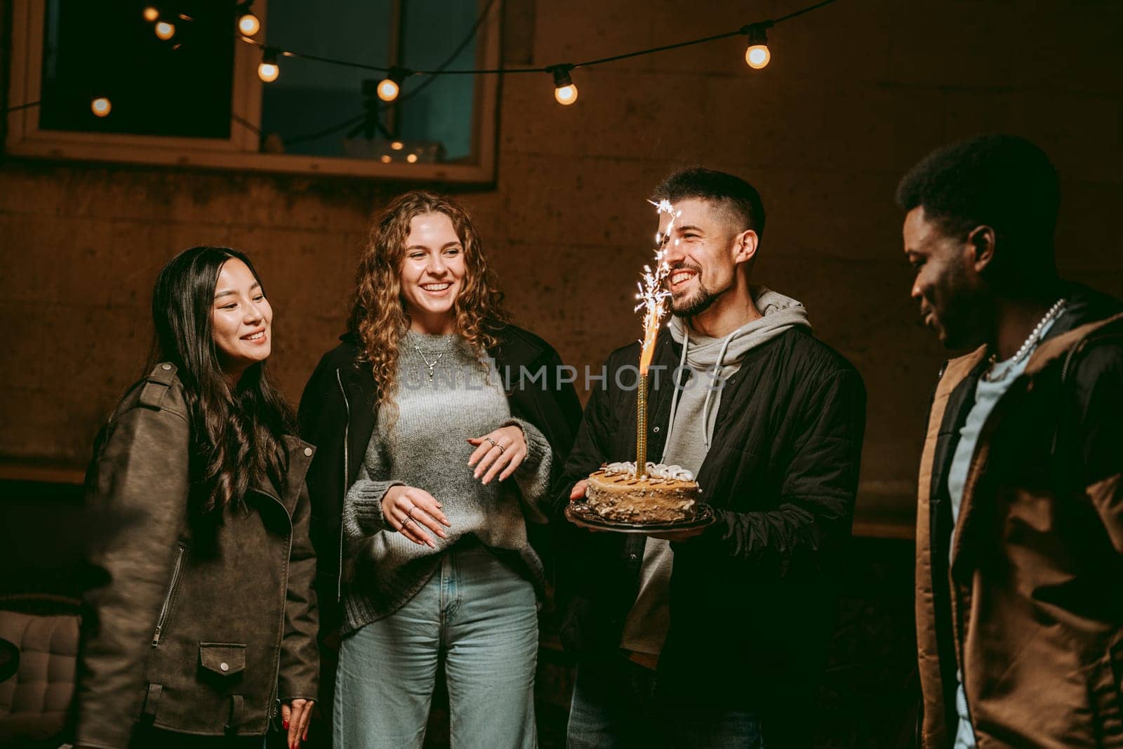 Group of friends gather outdoors at night to celebrate a birthday, sharing laughter and joy while enjoying cake topped with a sparkler. The atmosphere is festive and warm.