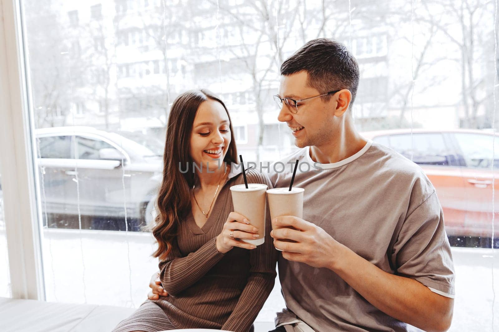 A couple sits closely by a large window in a cafe, smiling and holding warm drinks. Outside, snow is visible, enhancing the cozy atmosphere of their day together.