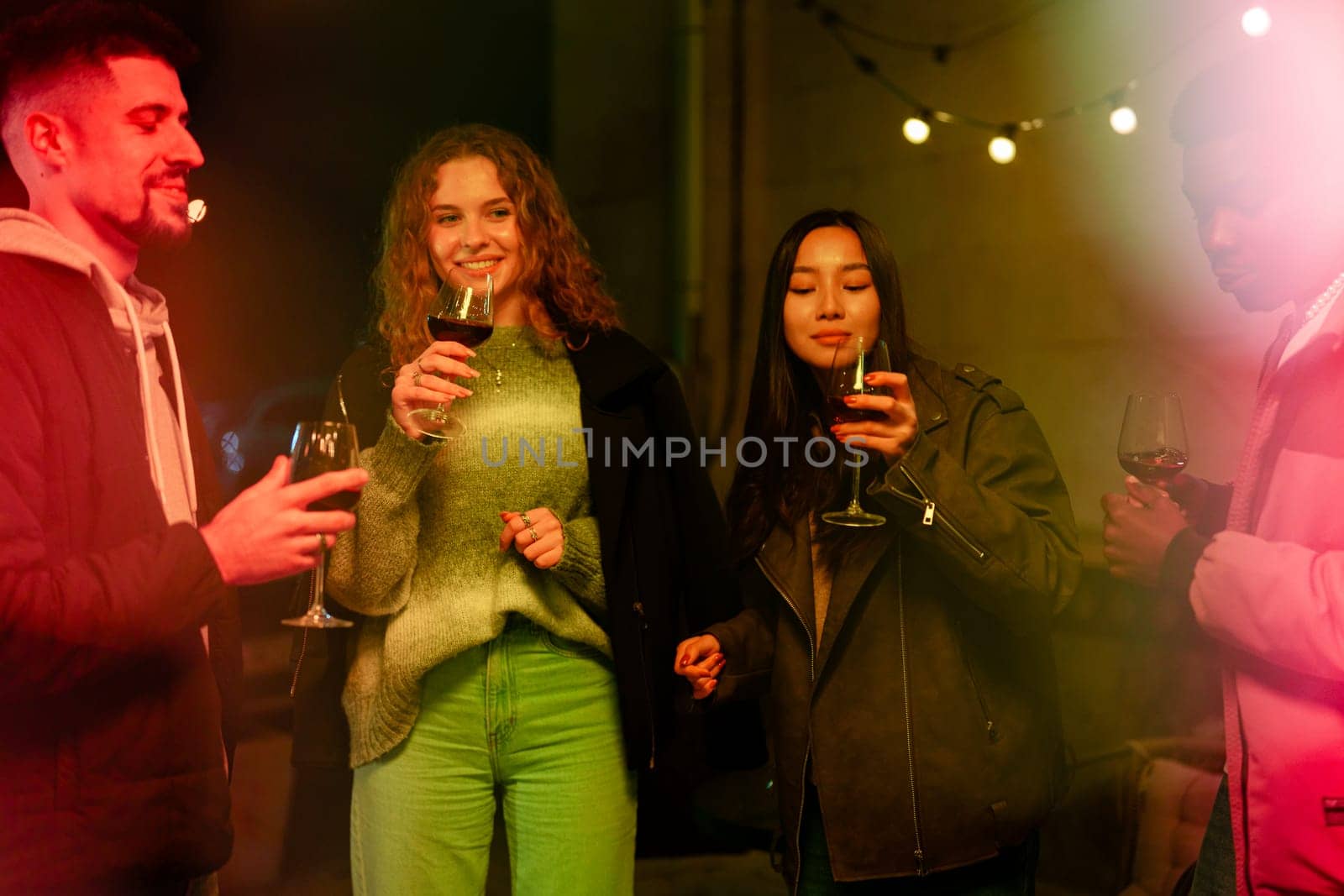 Friends gather in a cozy outdoor setting, raising their glasses filled with drinks while sharing laughter and conversation under warm string lights at night.