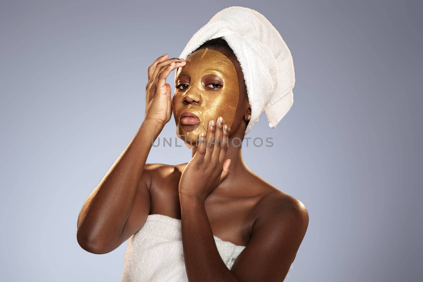 A woman stands in a studio applying a gold facial mask to her face. She wears a towel on her head and another towel wrapped around her body, enjoying a moment of self-care and relaxation.