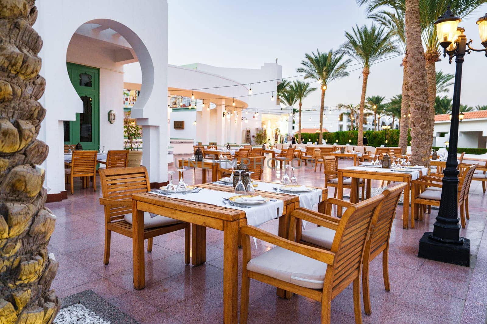 This setting features elegantly arranged wooden tables and chairs under a tranquil sky. Lush palm trees surround the outdoor dining space, creating a serene atmosphere.