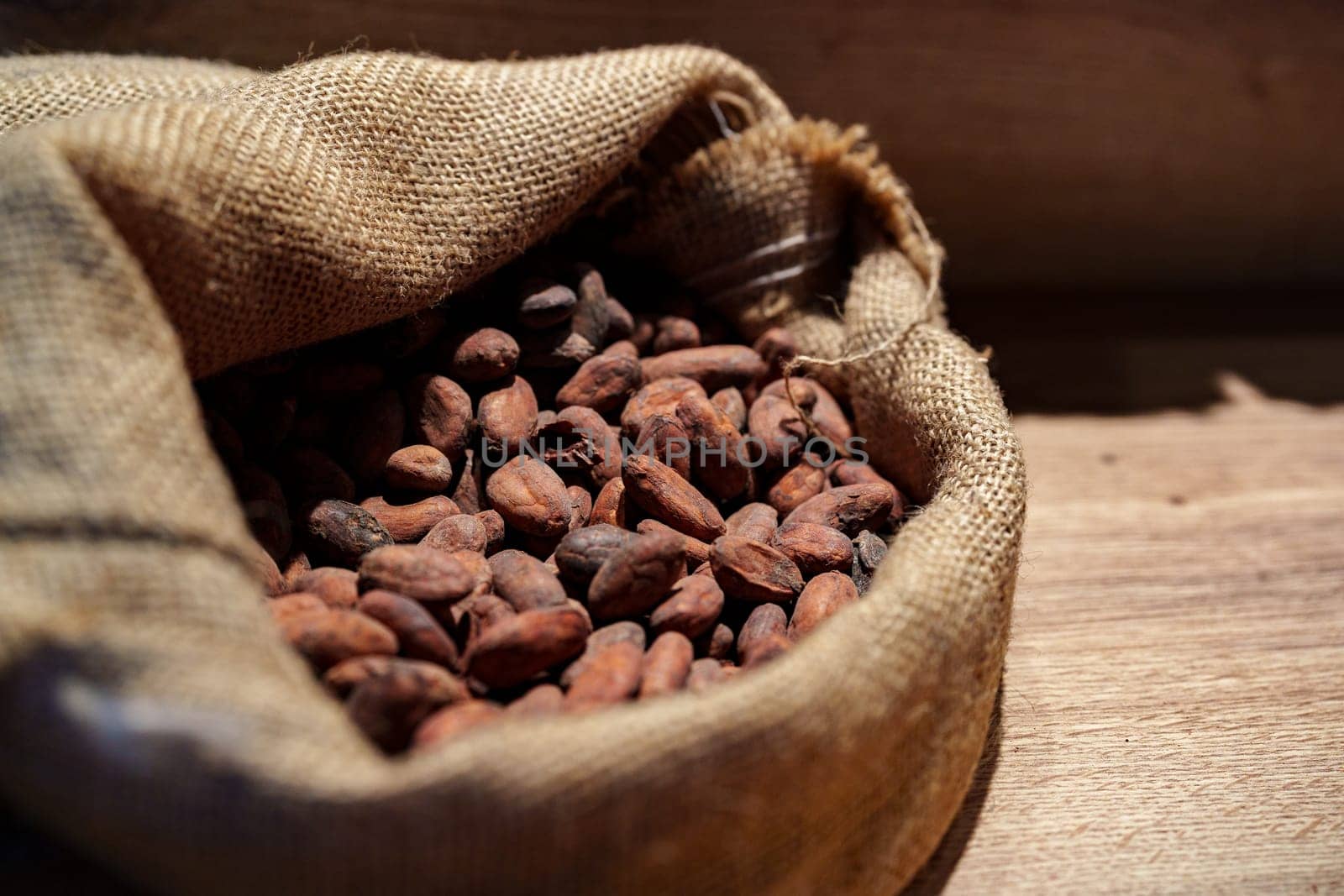 Cacao beans rest in a rustic burlap sack on a wooden table. The warm lighting highlights the rich colors of the beans, showcasing their natural beauty and texture.