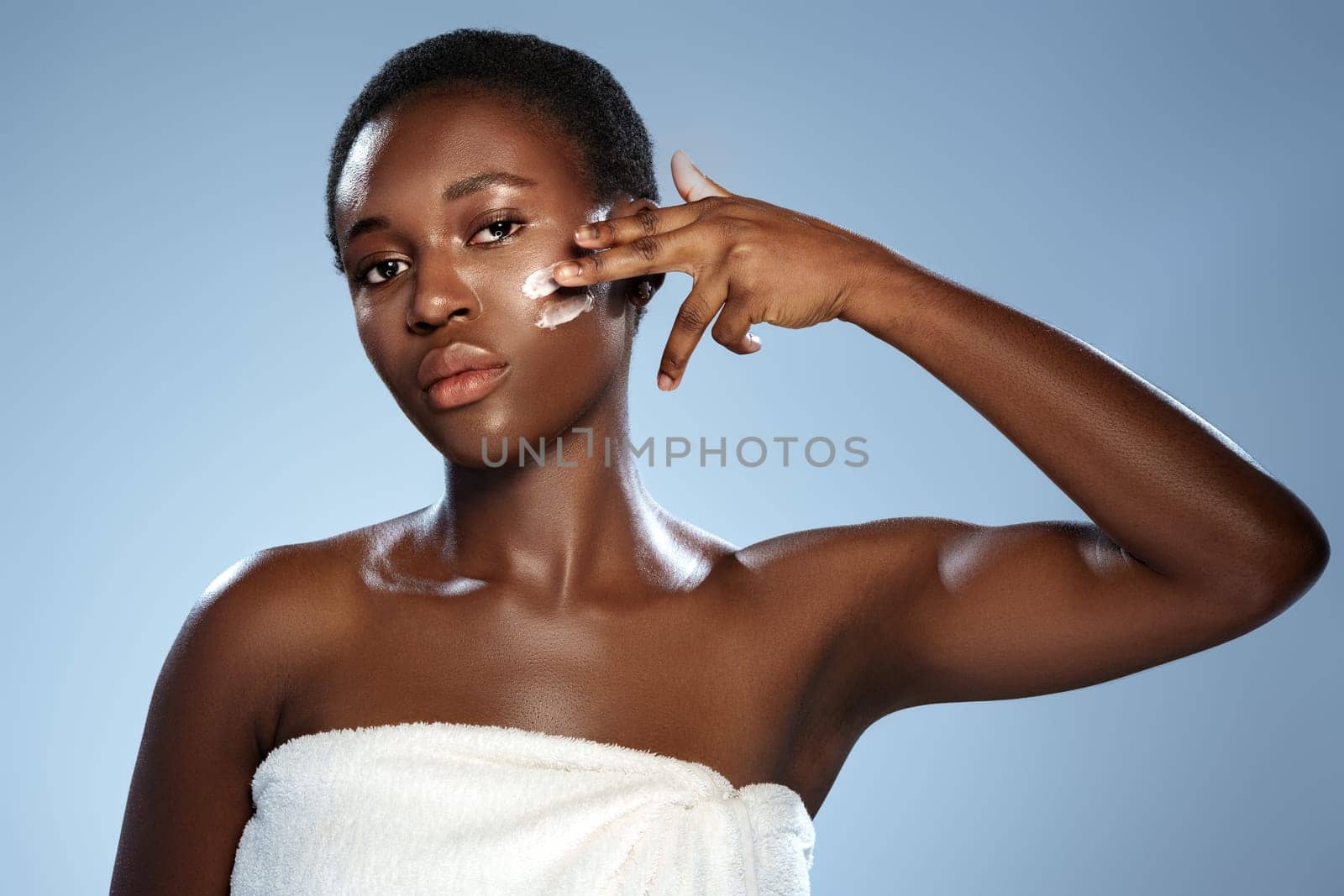 A woman is gently applying a skincare product to her face in a well-lit studio with a soft blue background. She looks focused and relaxed as she takes care of her skin.